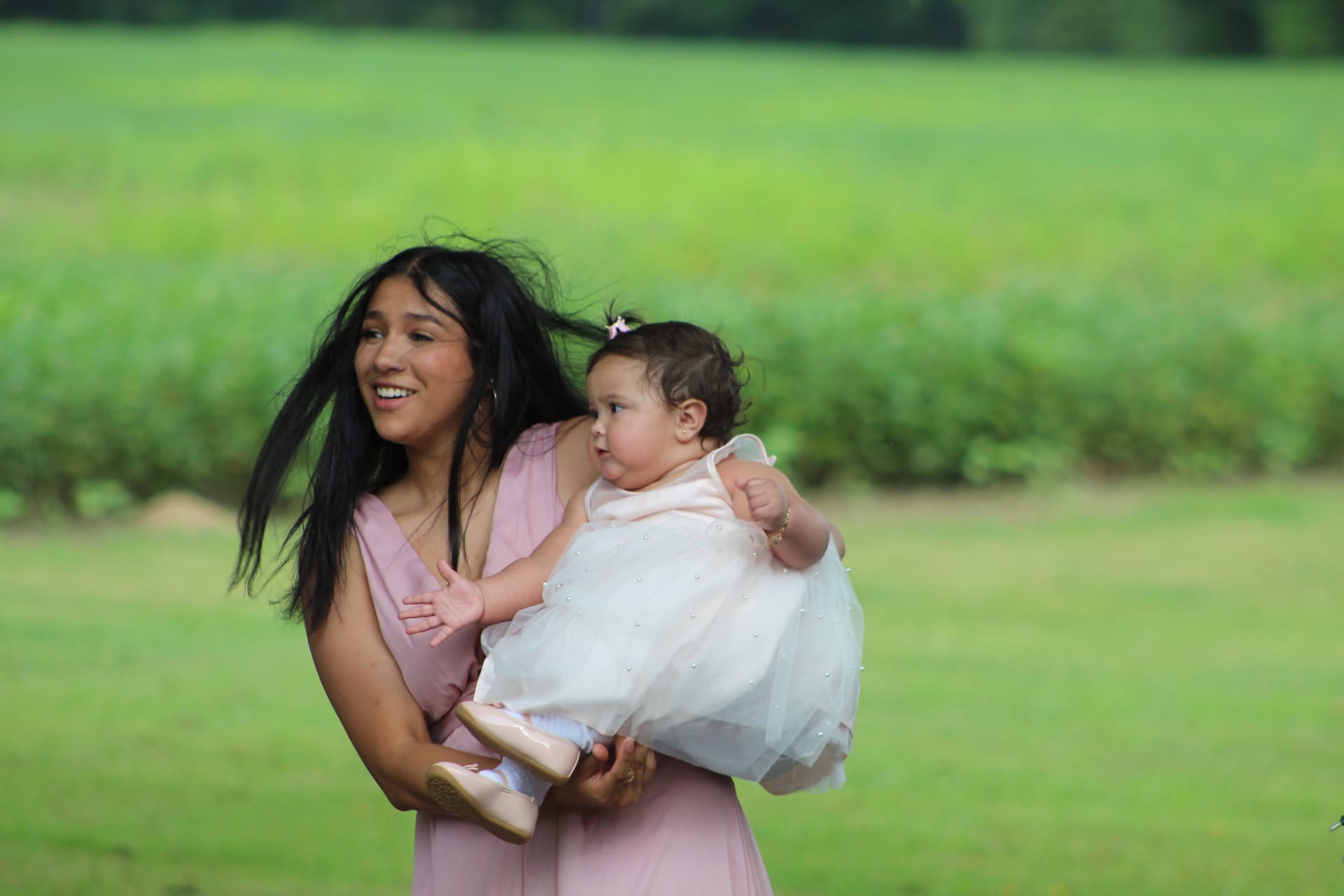 A smiling young woman in a blush pink gown holds a chubby baby dressed in a white tulle outfit on the open farmland fields of Scarborough House estate.