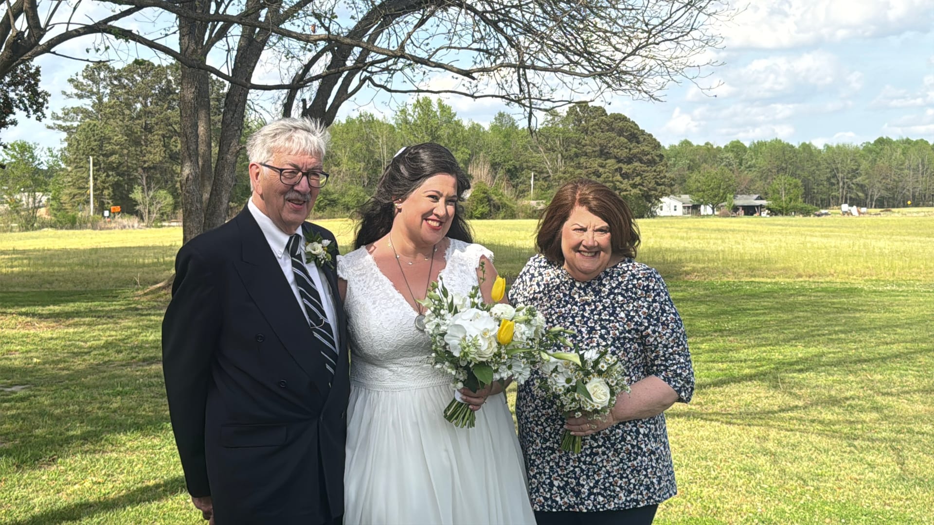 A smiling bride in a white gown holding a bouquet of white roses and yellow tulips stands between two proud family members on the sunlit grounds of a historic estate wedding venue.