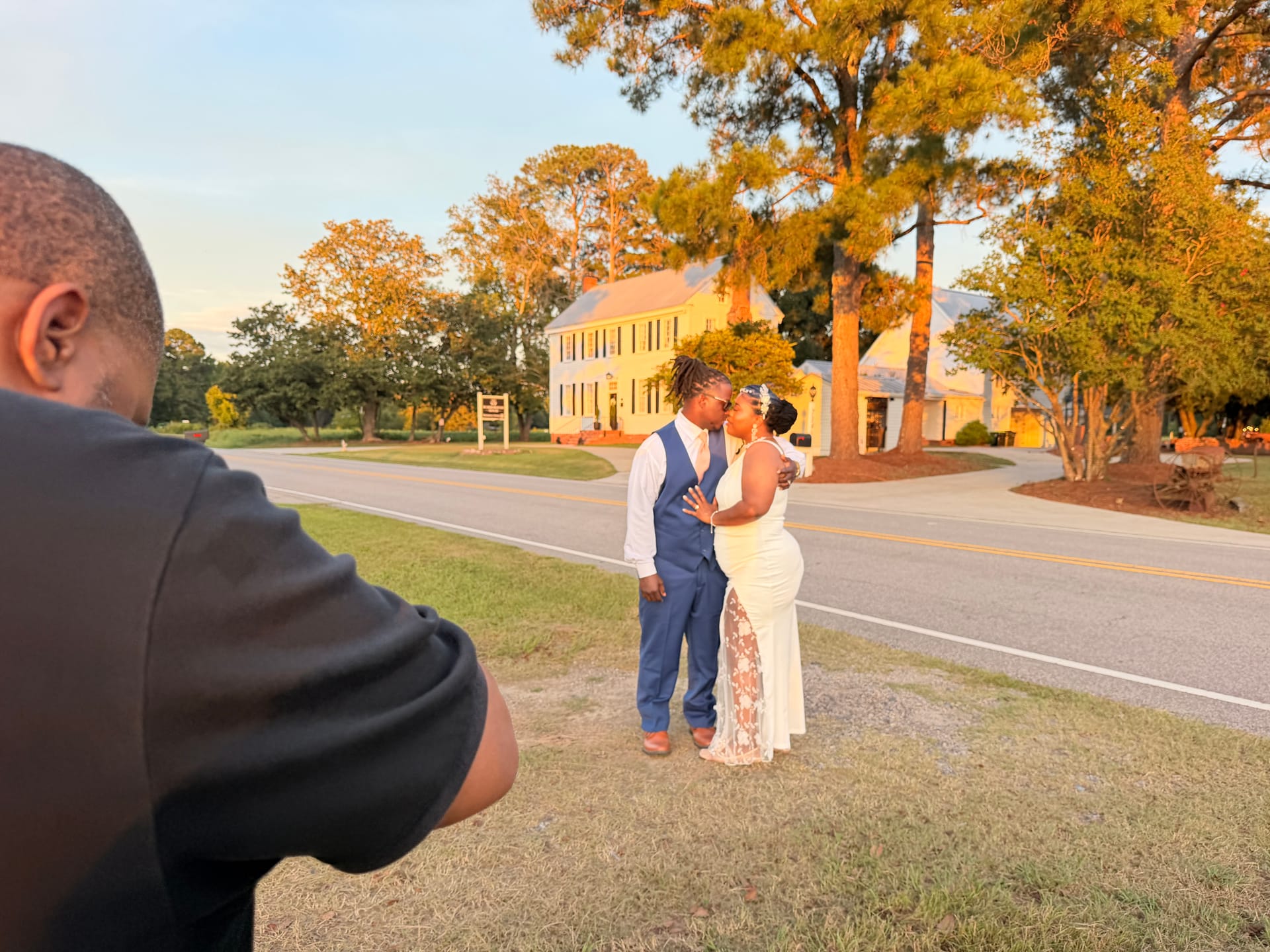 A couple shares a kiss on the lawn at golden hour in front of the 1821 Scarborough House manor while a photographer captures the moment.