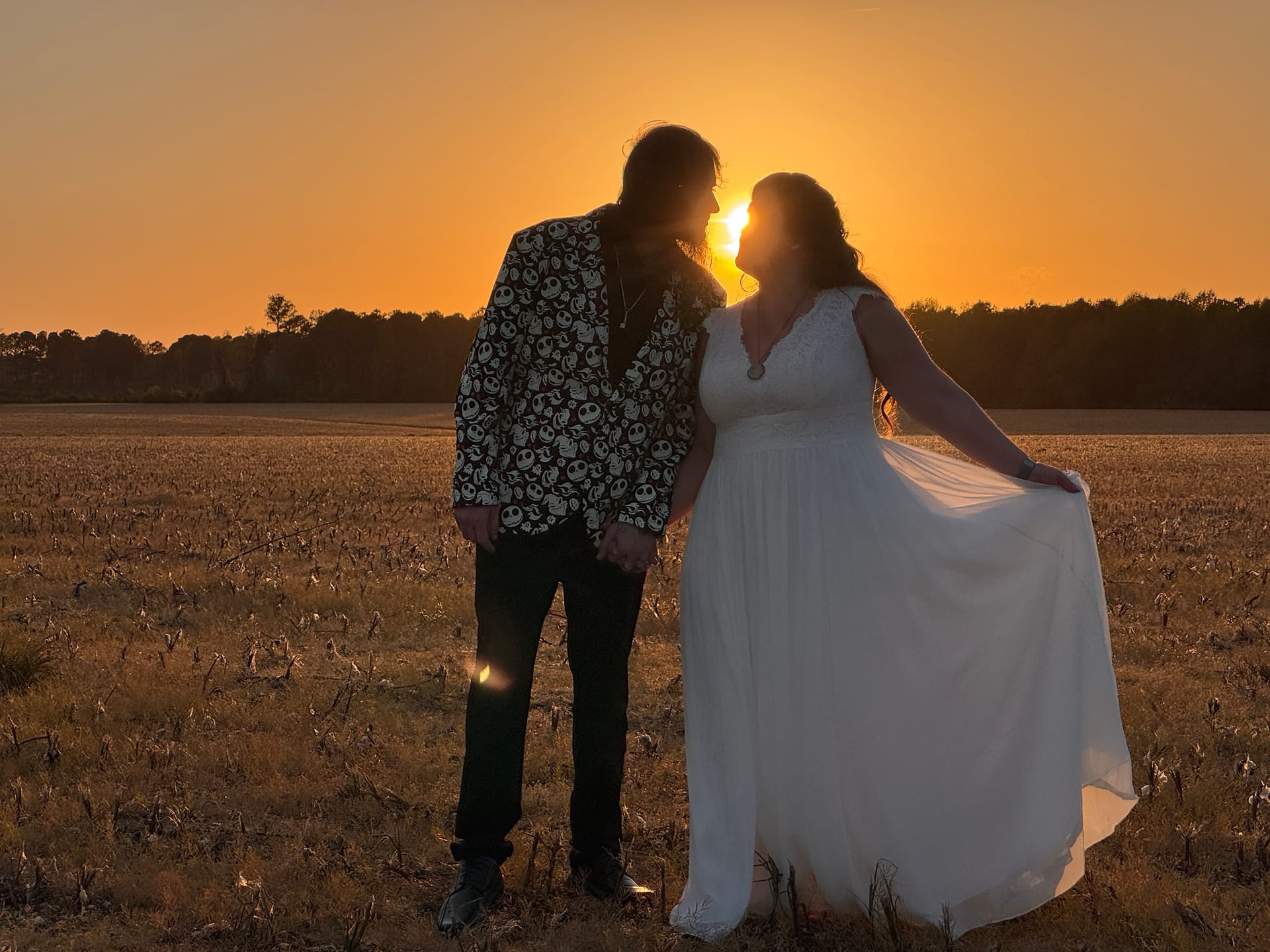 Bride in flowing white gown and groom in a patterned blazer share a romantic moment face-to-face against a vibrant golden sunset over open farmland fields at Scarborough House.