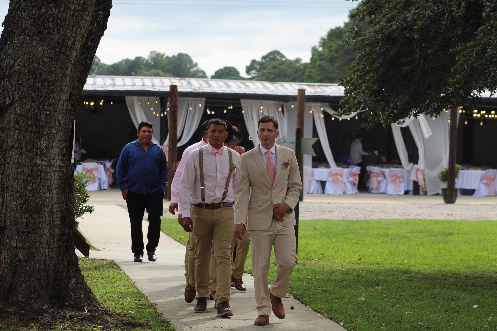 Groomsmen in tan suits and pink bow ties walk along a tree-lined pathway toward the camera at a rustic outdoor wedding venue with draped white fabric and string lights in the background.