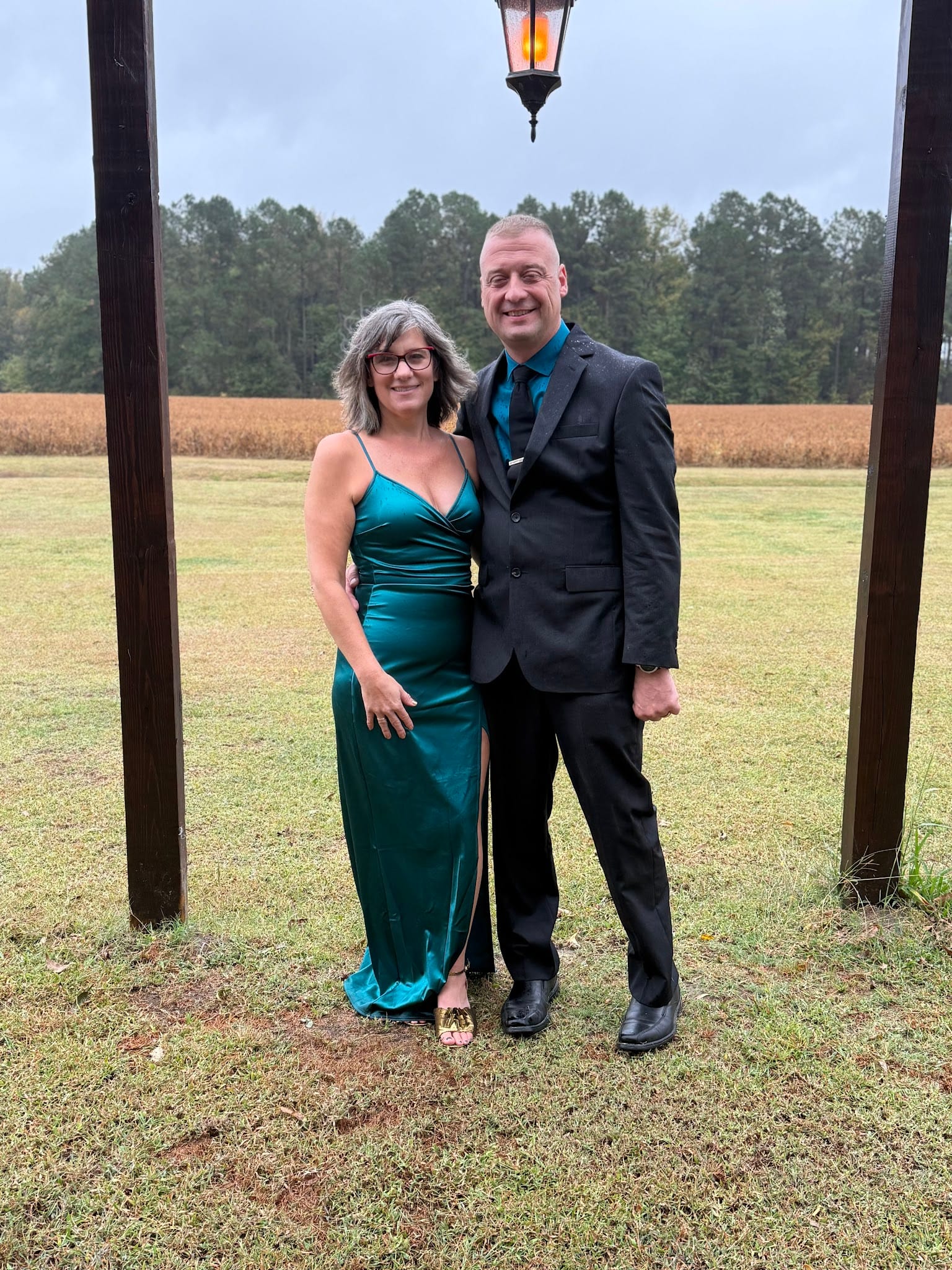 Elegantly dressed couple posing under a hanging lantern between wooden posts with golden farmland fields and pine trees in the background at Scarborough House.