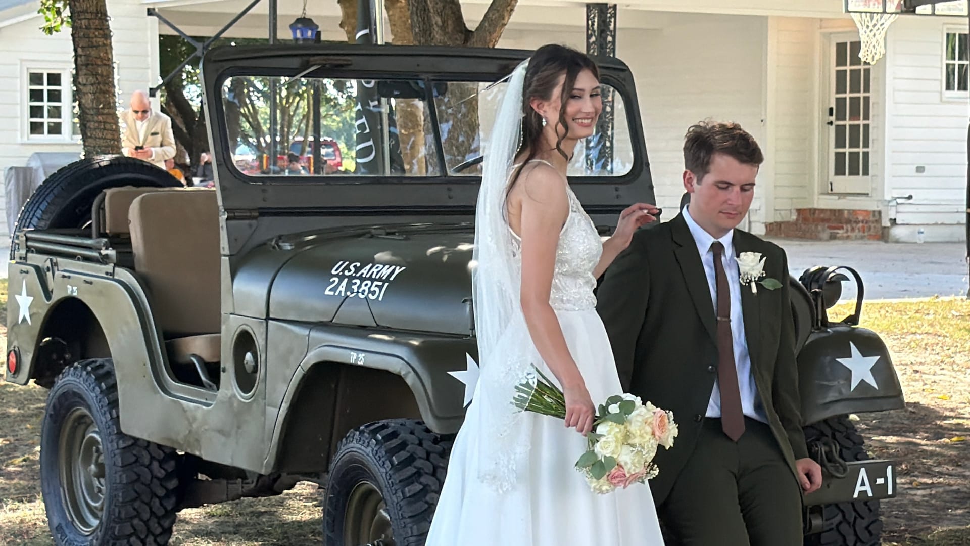 Bride in a white gown holding a bouquet smiles beside her groom in an olive suit as they pose in front of a vintage U.S. Army Jeep on the grounds of Scarborough House historic estate.
