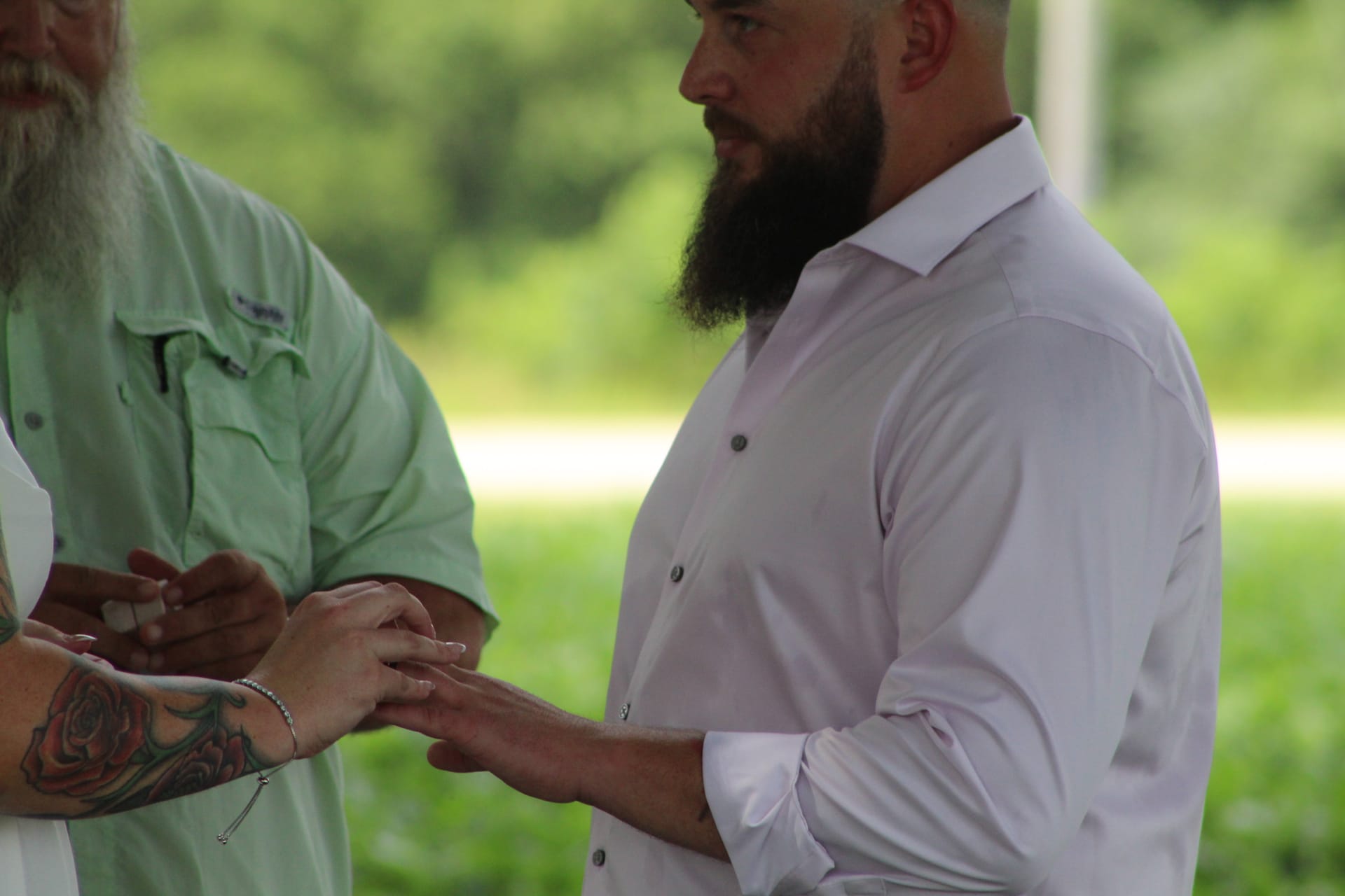 Bearded groom in a white dress shirt places a wedding ring on his brides tattooed hand during an outdoor ceremony at Scarborough House.