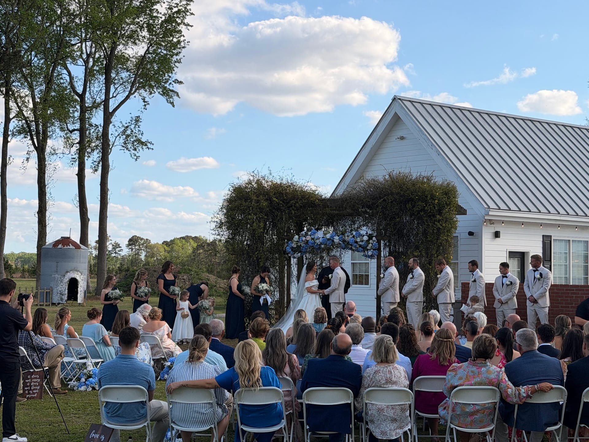 Guests seated on white chairs watch a wedding ceremony at Scarborough House as the couple exchanges vows beneath a lush greenery and blue floral arch beside a white farmhouse chapel on a sunny day.