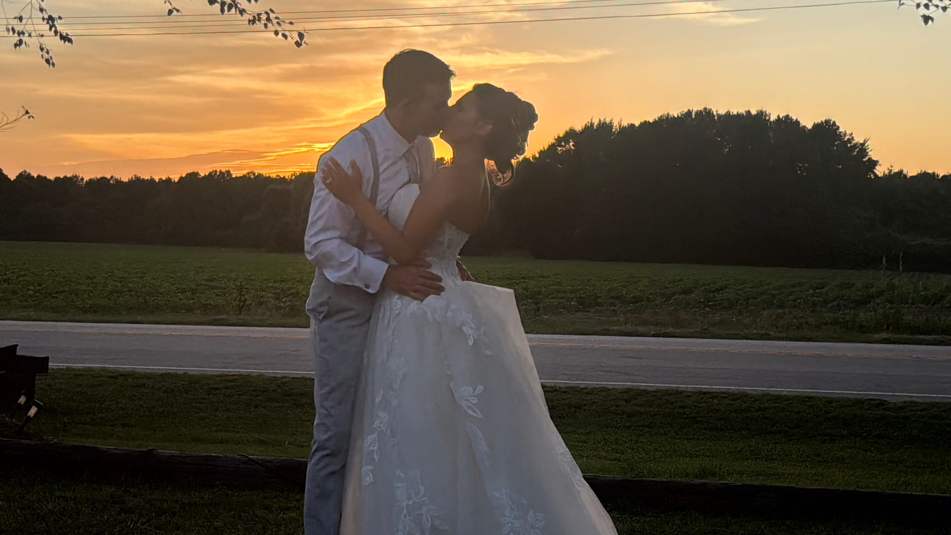 Bride in a floral lace ball gown and groom in a white shirt with suspenders share a kiss against a glowing golden sunset sky overlooking open farmland fields at Scarborough House.