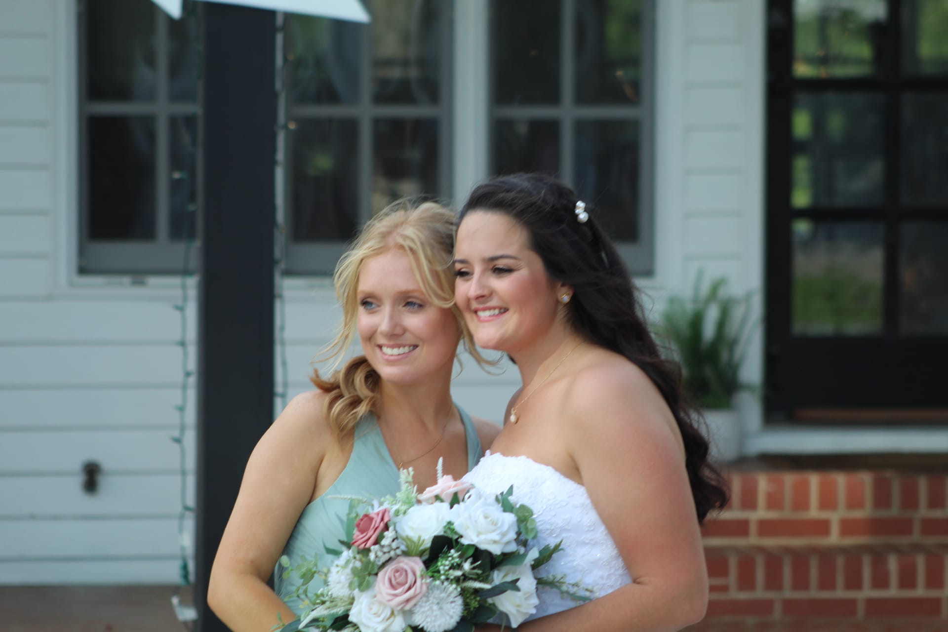 Bride in strapless white gown holding a blush and white rose bouquet laughs with her bridesmaid in a sage green dress in front of the historic 1821 Scarborough House manor.