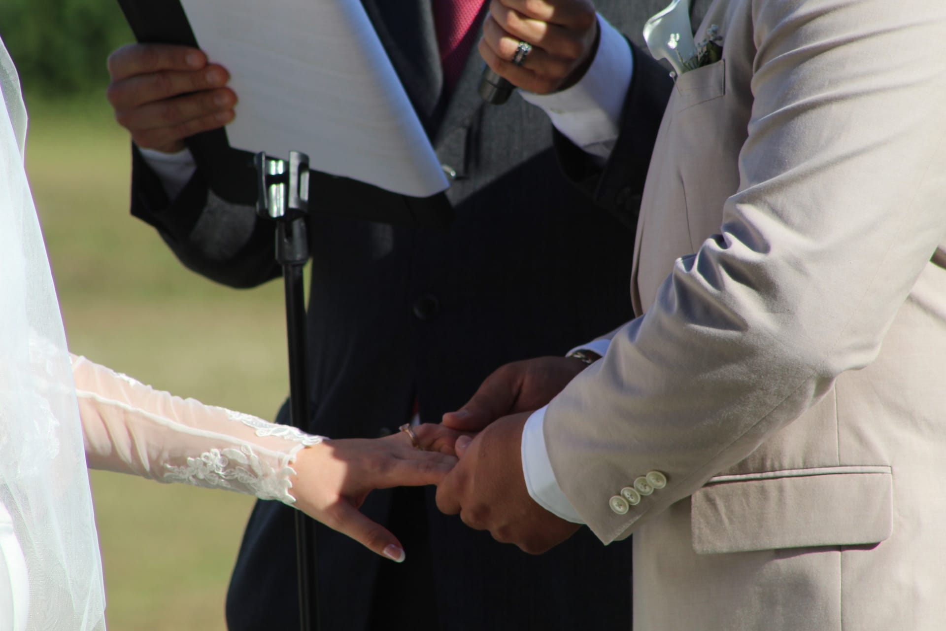 Groom in a tan suit places a wedding ring on the brides lace-sleeved hand during an outdoor ceremony at Scarborough House while the officiant reads from a book in the background.
