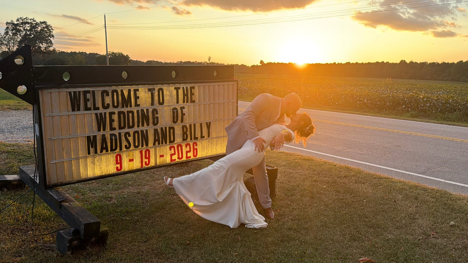 Bride and groom share a romantic dip kiss in front of a glowing marquee wedding welcome sign at golden hour with a fiery sunset over open farmland fields.