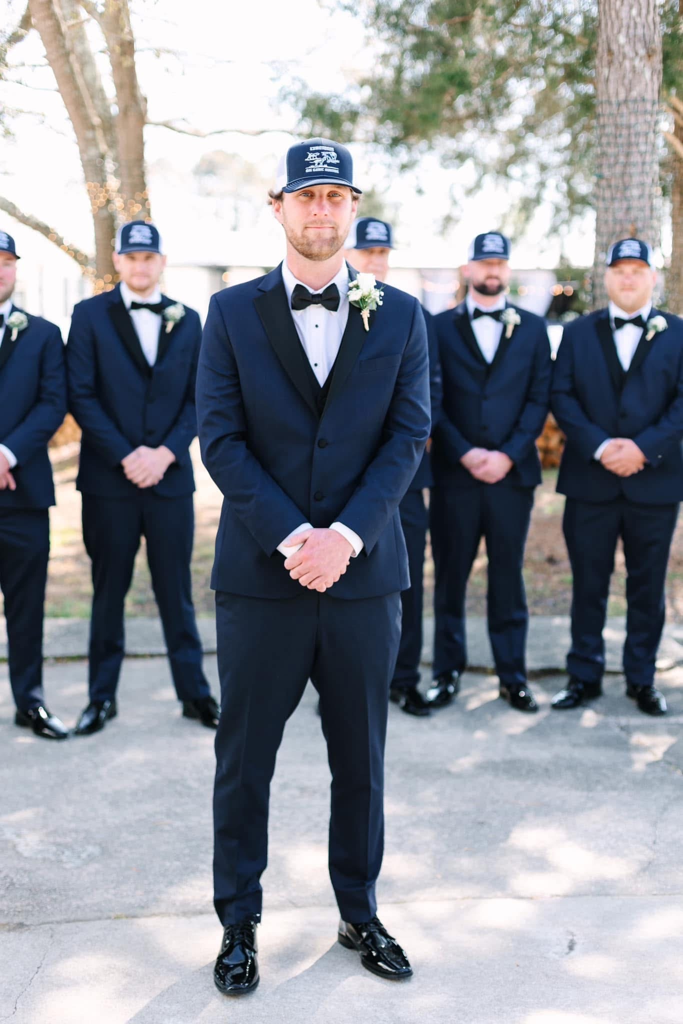 Groom in navy tuxedo and boutonniere stands in front of his groomsmen all wearing matching navy suits and coordinating baseball caps on the grounds of Scarborough House wedding venue.