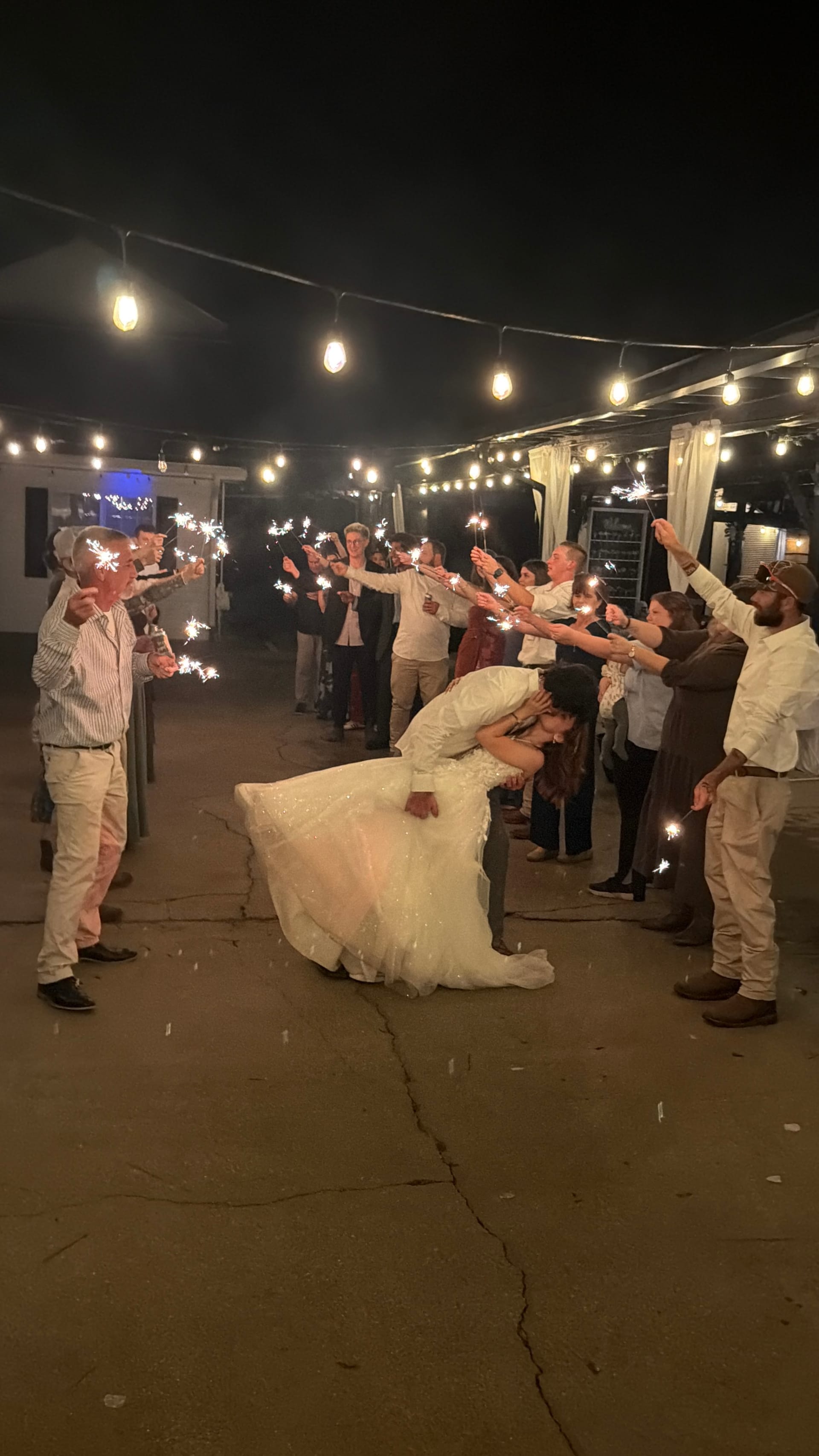 Bride and groom share a romantic dip kiss during their sparkler exit at night surrounded by cheering guests holding sparklers under warm string lights at Scarborough House.