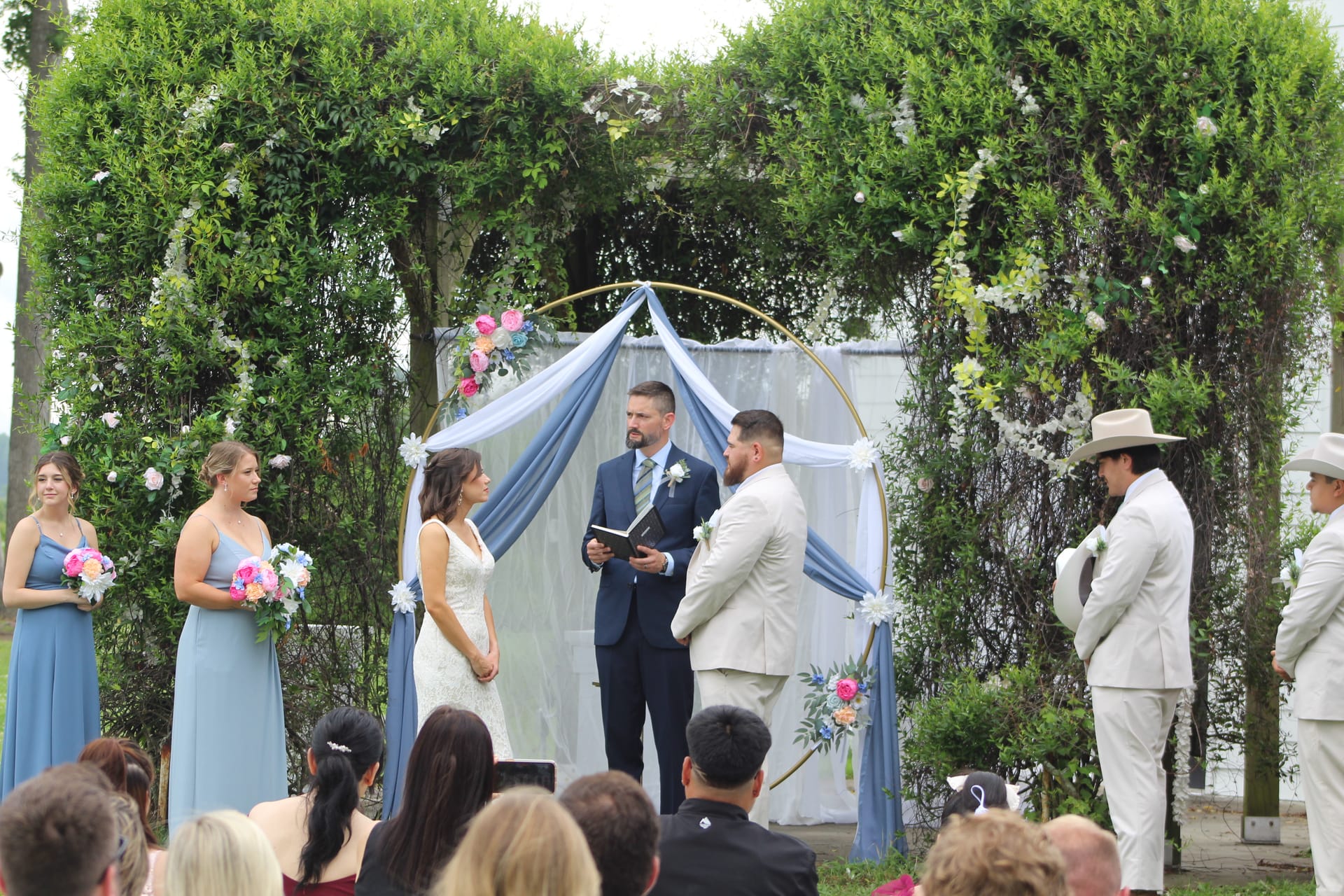 Bride and groom exchange vows beneath a draped floral hoop arch surrounded by lush greenery at Scarborough House outdoor wedding ceremony.