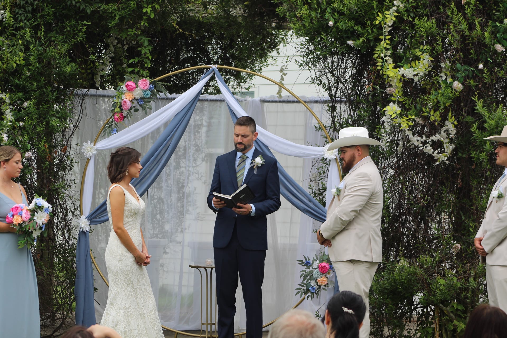 Bride and groom exchange vows beneath a gold circular arch adorned with dusty blue draping and colorful florals during an outdoor wedding ceremony at Scarborough House.