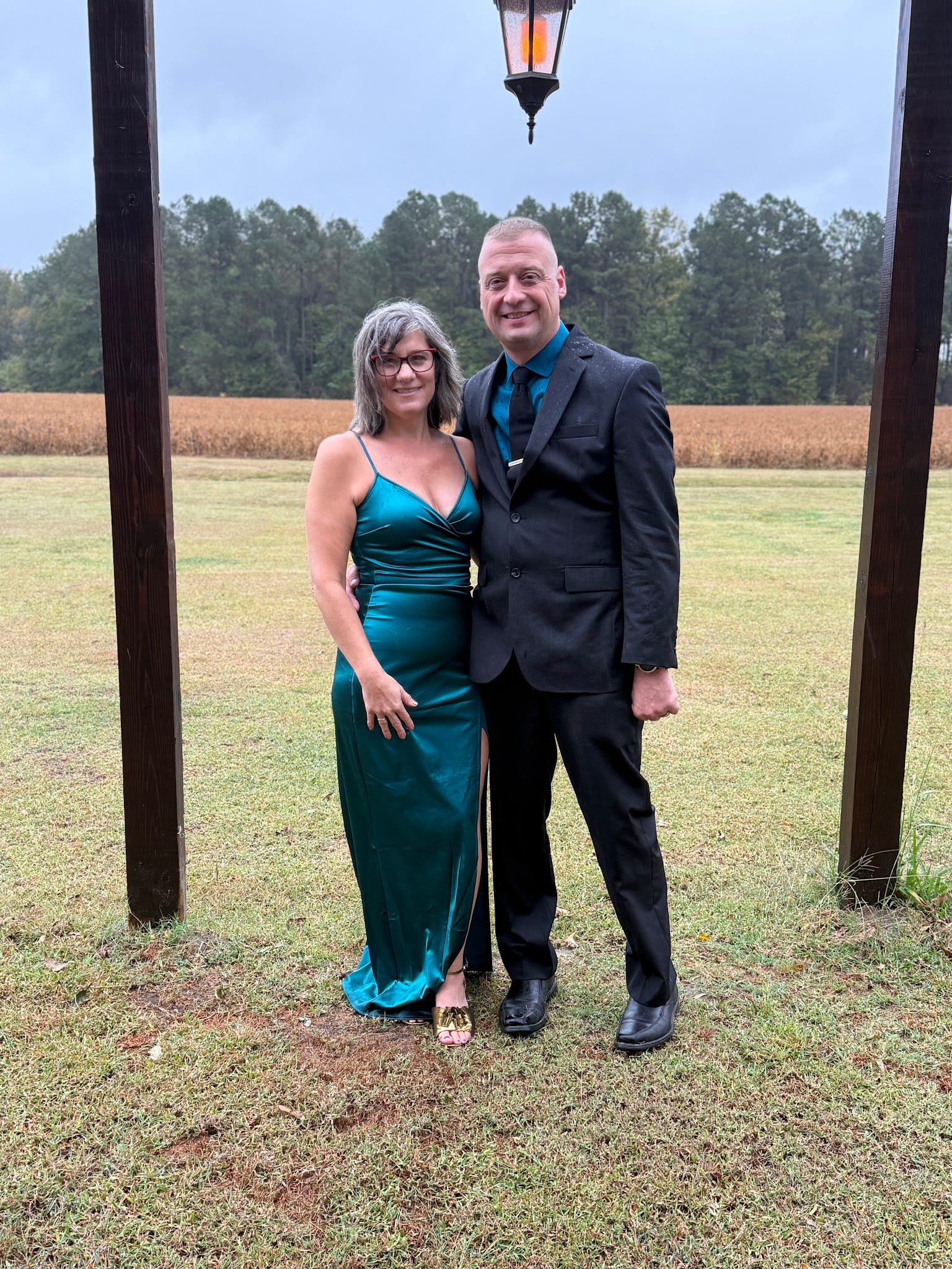 Elegantly dressed couple posing under a rustic lantern structure with open farmland fields and pine trees in the background at Scarborough House wedding venue.