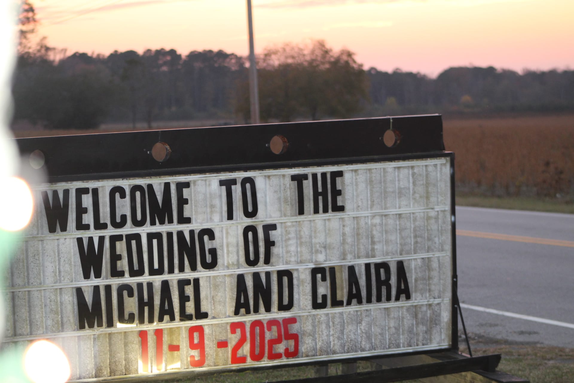 A roadside marquee sign welcomes guests to the wedding of Michael and Claira on November 9 2025 against a pastel sunset sky with farmland fields in the background.