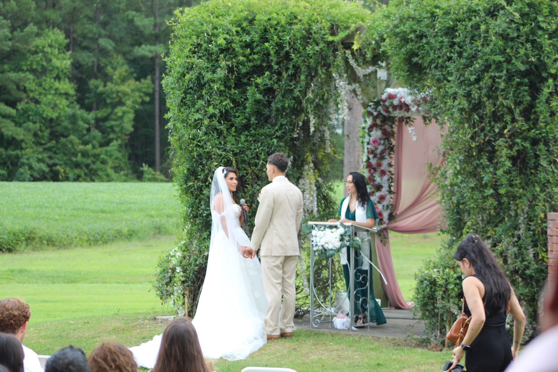 Bride and groom exchange vows beneath a lush ivy-covered arch adorned with floral arrangements and blush draping on the ceremony lawn at Scarborough House estate.