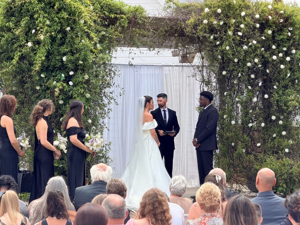 Bride and groom exchange vows beneath a lush green floral arch adorned with white roses and flowing white draping on the ceremony lawn at Scarborough House, surrounded by bridesmaids in black and seated wedding guests.