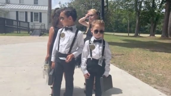 Three children dressed in black and white formal attire with bow ties suspenders and sunglasses carrying small briefcases on a sunlit pathway at Scarborough House wedding venue.