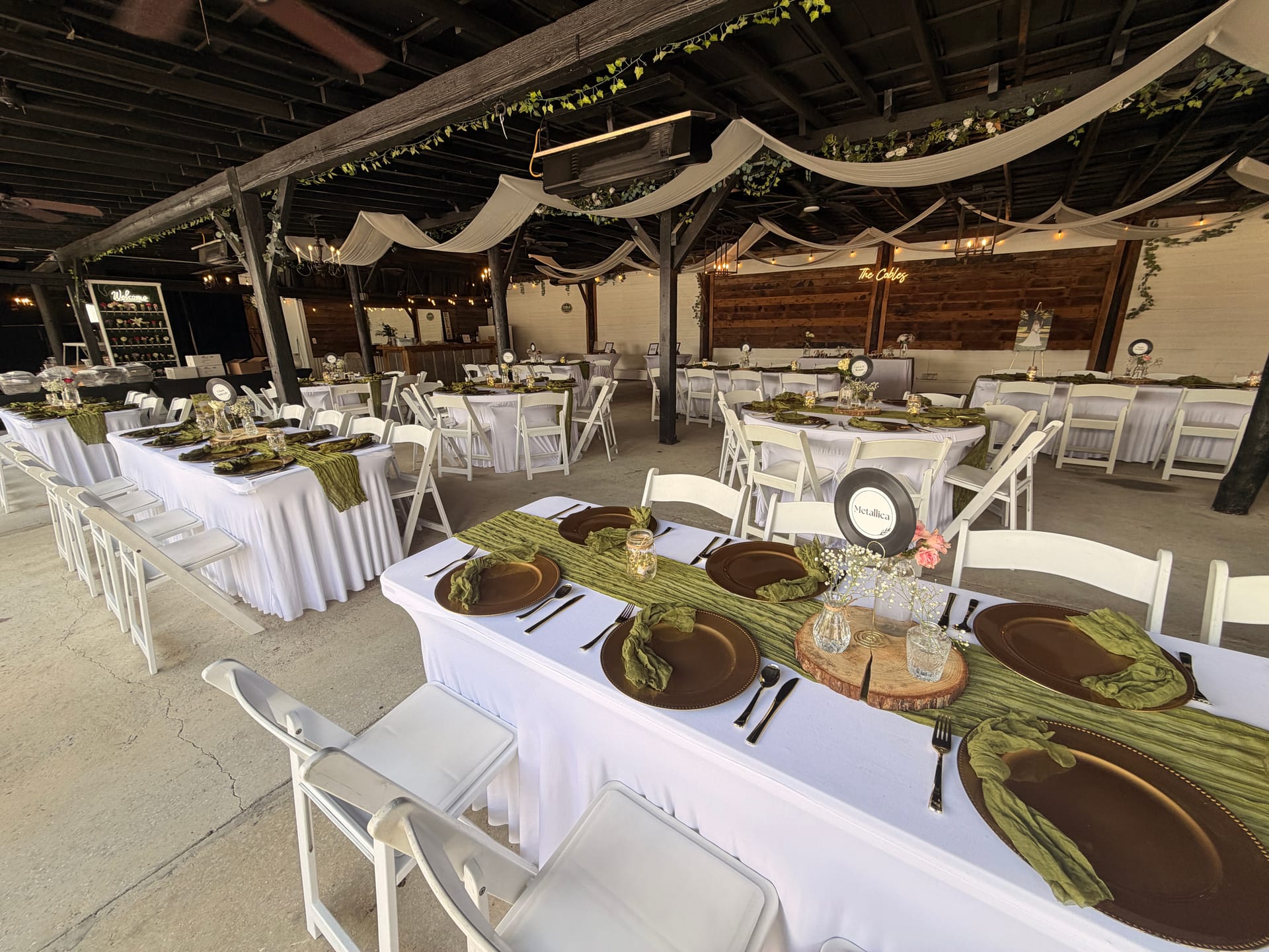 Elegantly set reception tables with white linens, copper charger plates, sage green runners, and floral centerpieces beneath draped fabric and string lights in a rustic open-air pavilion.