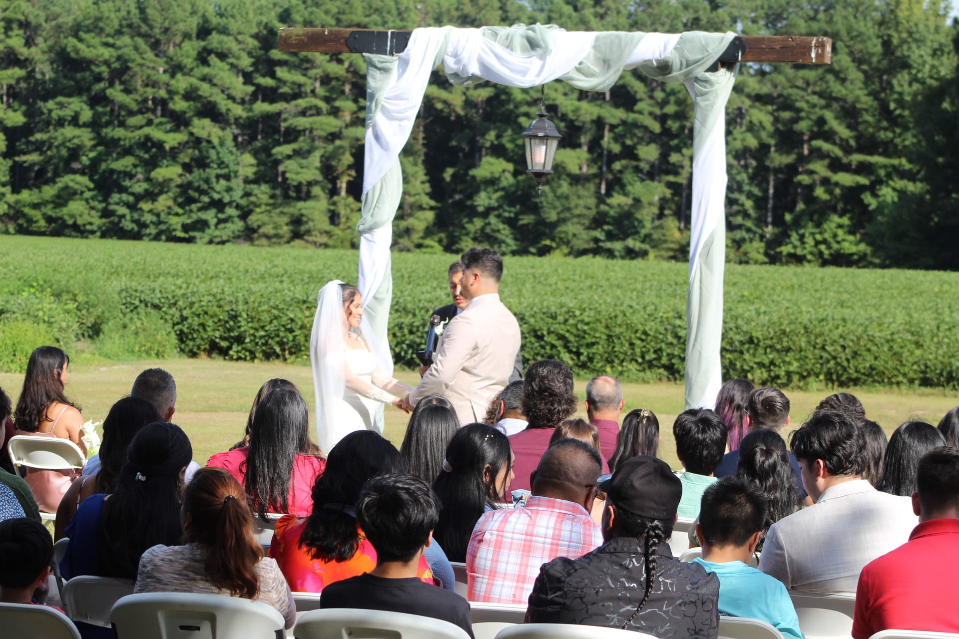 Bride and groom exchanging vows beneath a white-draped wooden arch on the ceremony lawn at Scarborough House, surrounded by seated guests with green farmland fields and trees in the background.