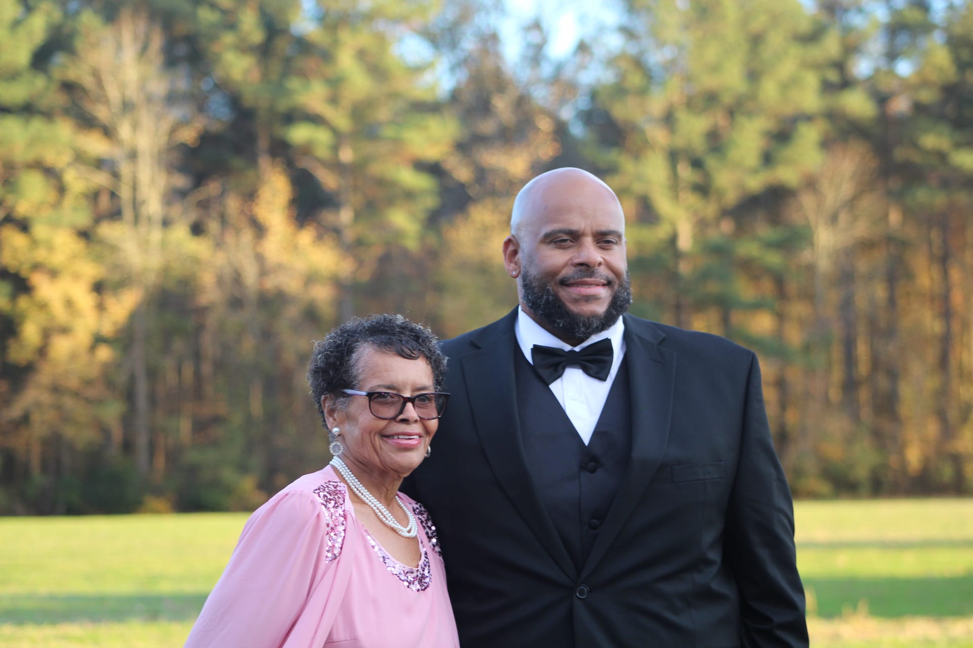A formally dressed man in a black tuxedo and bow tie poses with an older woman in a pink sequined dress and pearl necklace on the scenic grounds of a historic estate wedding venue surrounded by autumn trees.