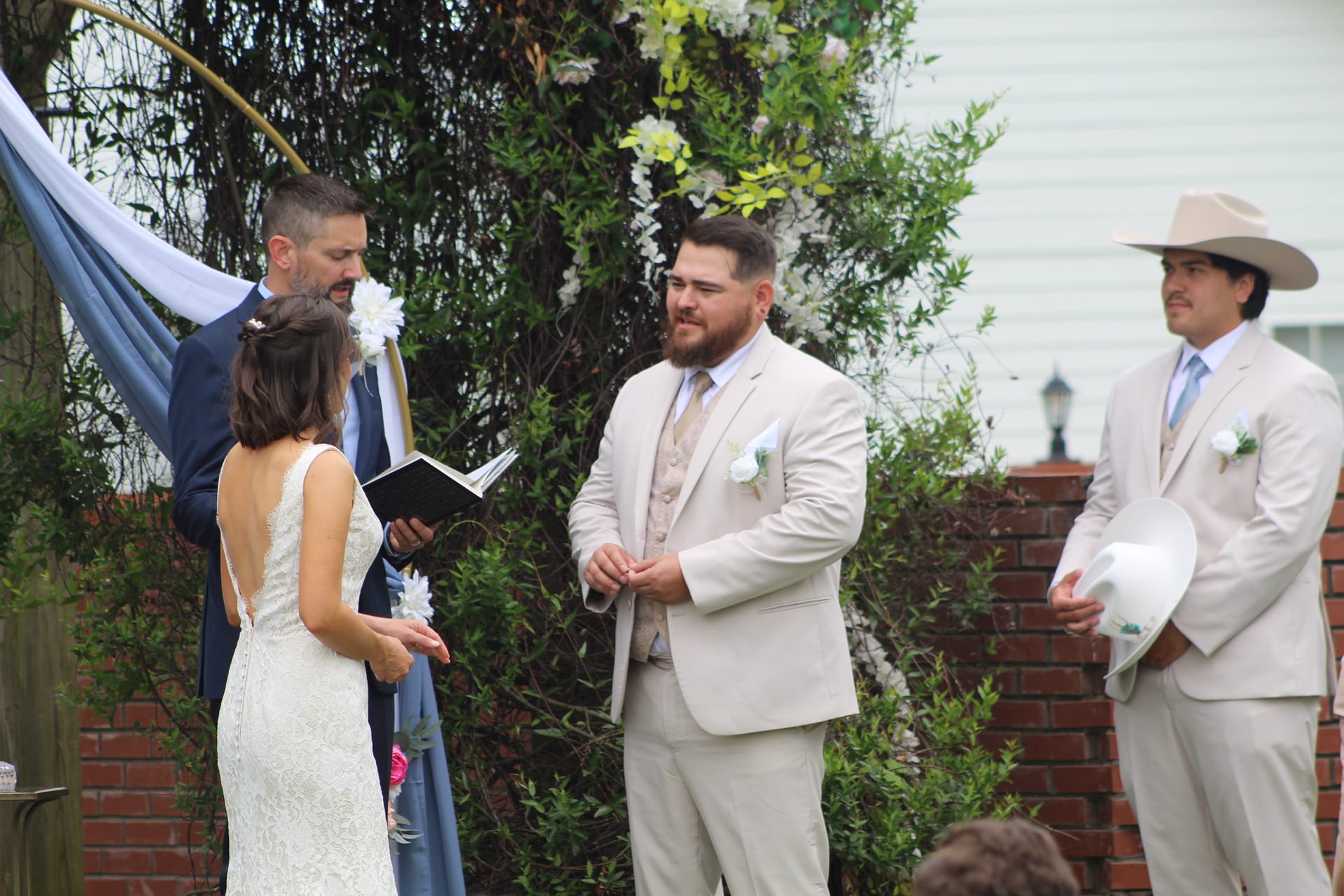 Groom in a tan suit exchanges vows with his bride beneath a floral-adorned circular arch on the ceremony lawn at Scarborough House while the officiant reads from a book and a groomsman stands nearby.