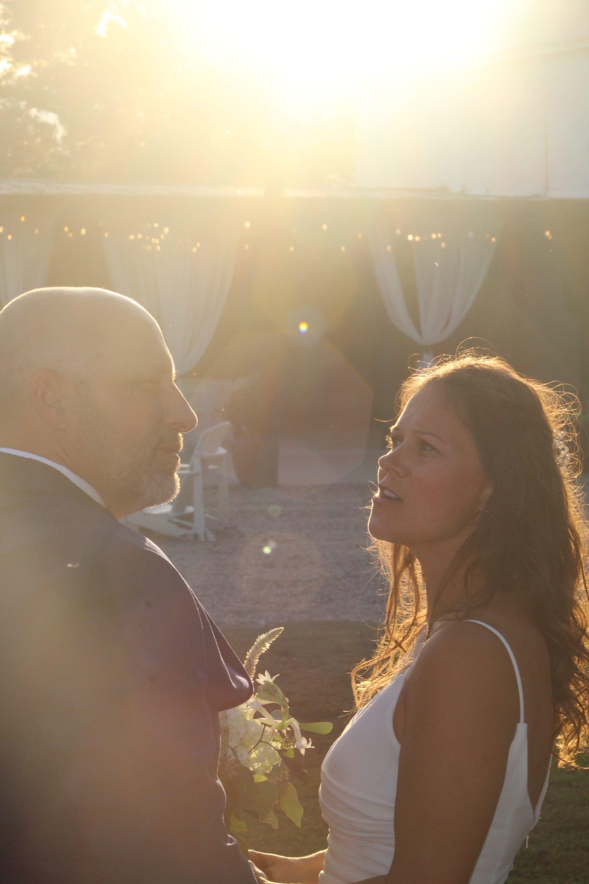 Bride in white dress gazes lovingly up at groom during outdoor wedding ceremony bathed in warm golden sunset light at Scarborough House estate.