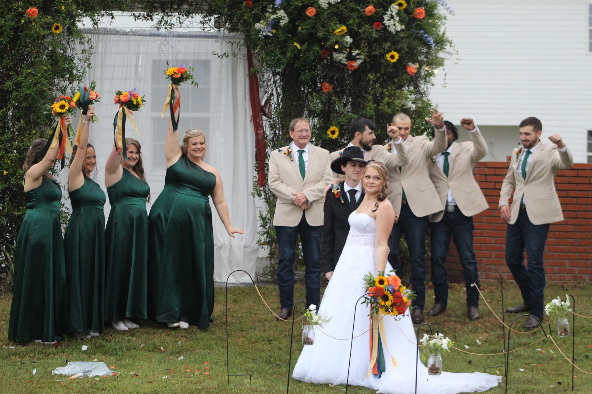 Bride and groom pose in front of a floral arch at Scarborough House while bridesmaids in emerald gowns raise colorful bouquets and groomsmen in tan blazers cheer behind them on the ceremony lawn.
