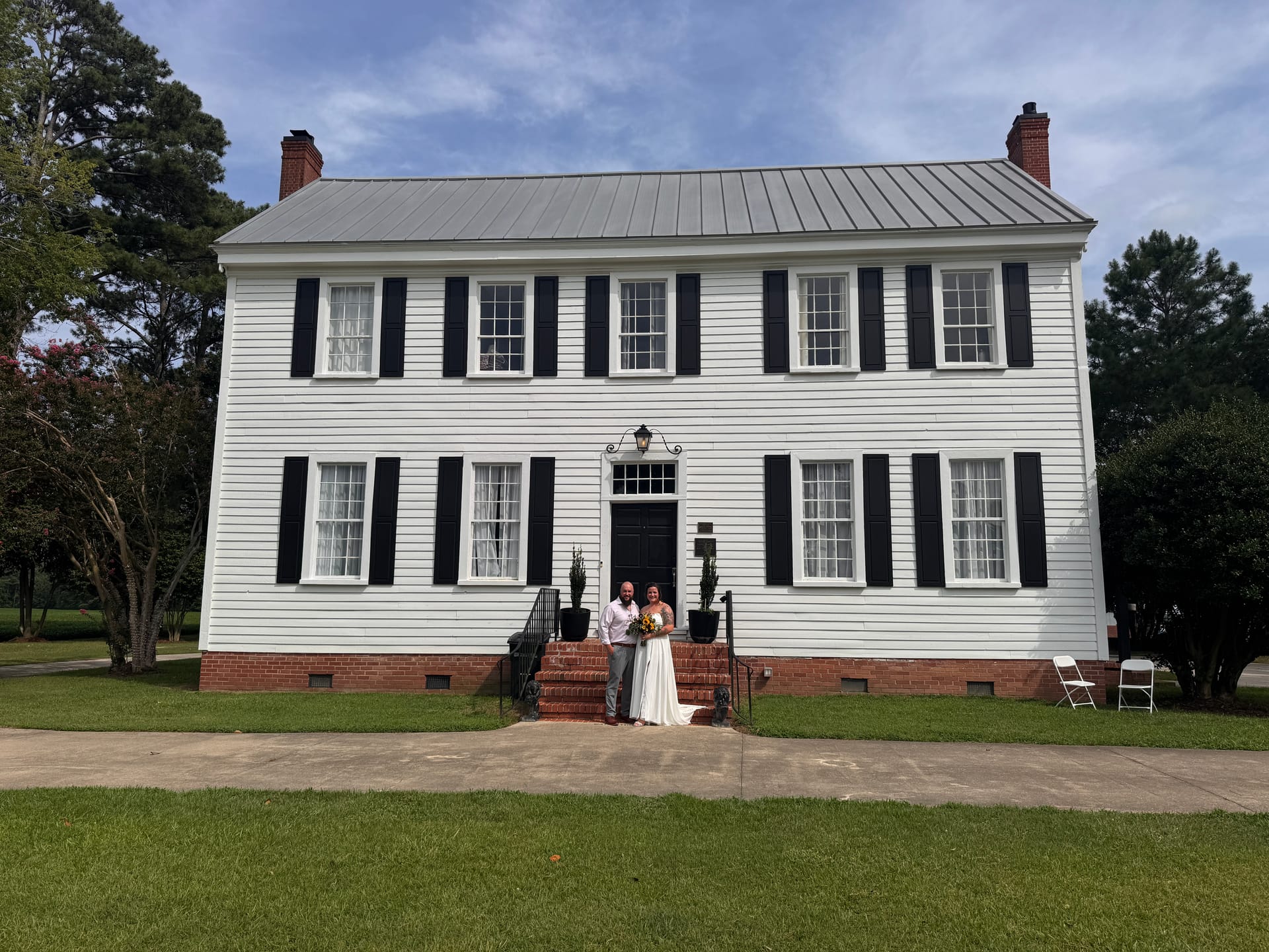 Bride and groom stand together on the brick front steps of the white 1821 Scarborough House manor, framed by black shutters, twin topiaries, and a metal roof under a partly cloudy sky.