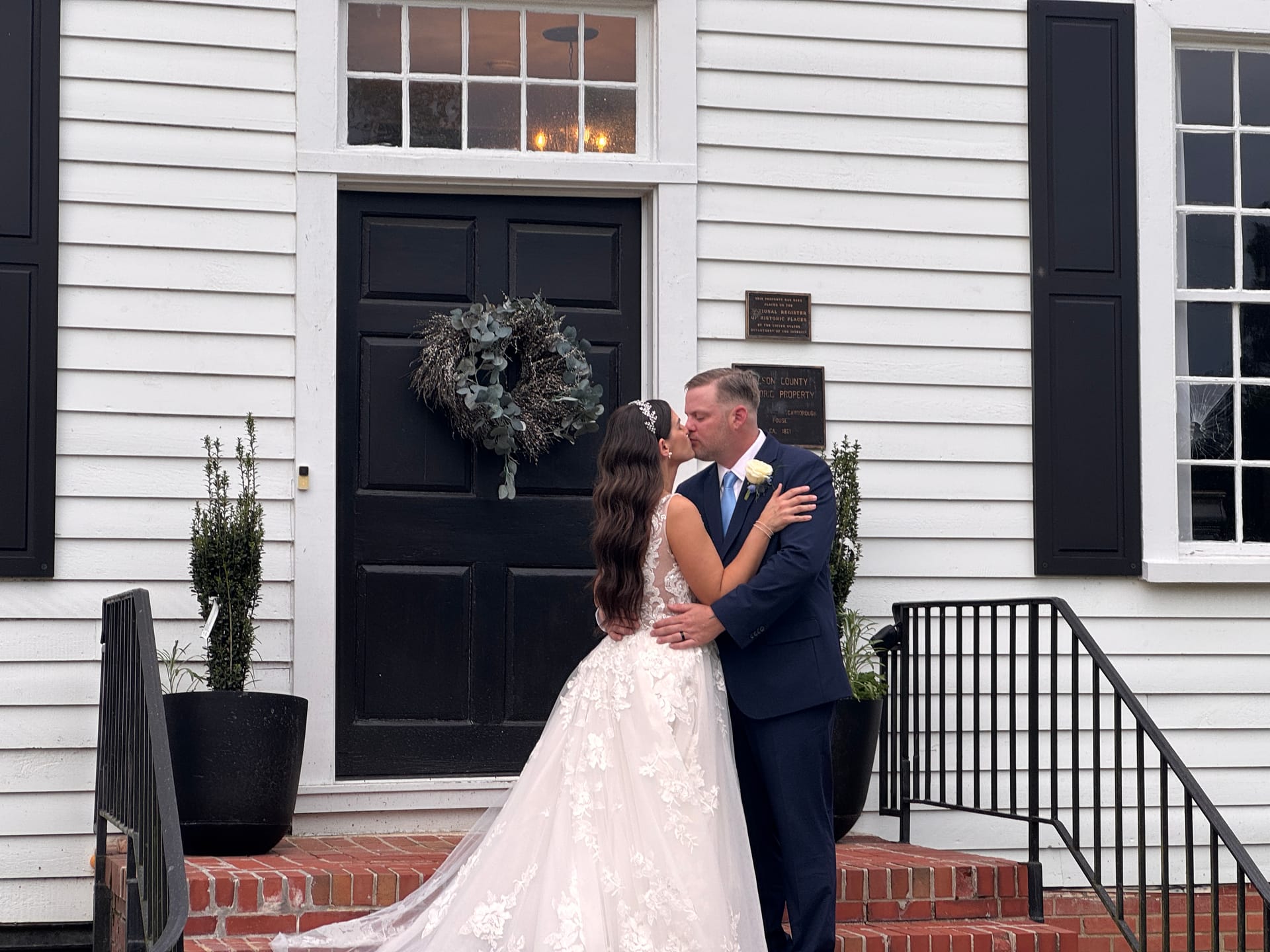 Bride and groom share their first kiss on the brick front steps of the 1821 Scarborough House manor, framed by a eucalyptus wreath on the black door and lush potted topiaries.