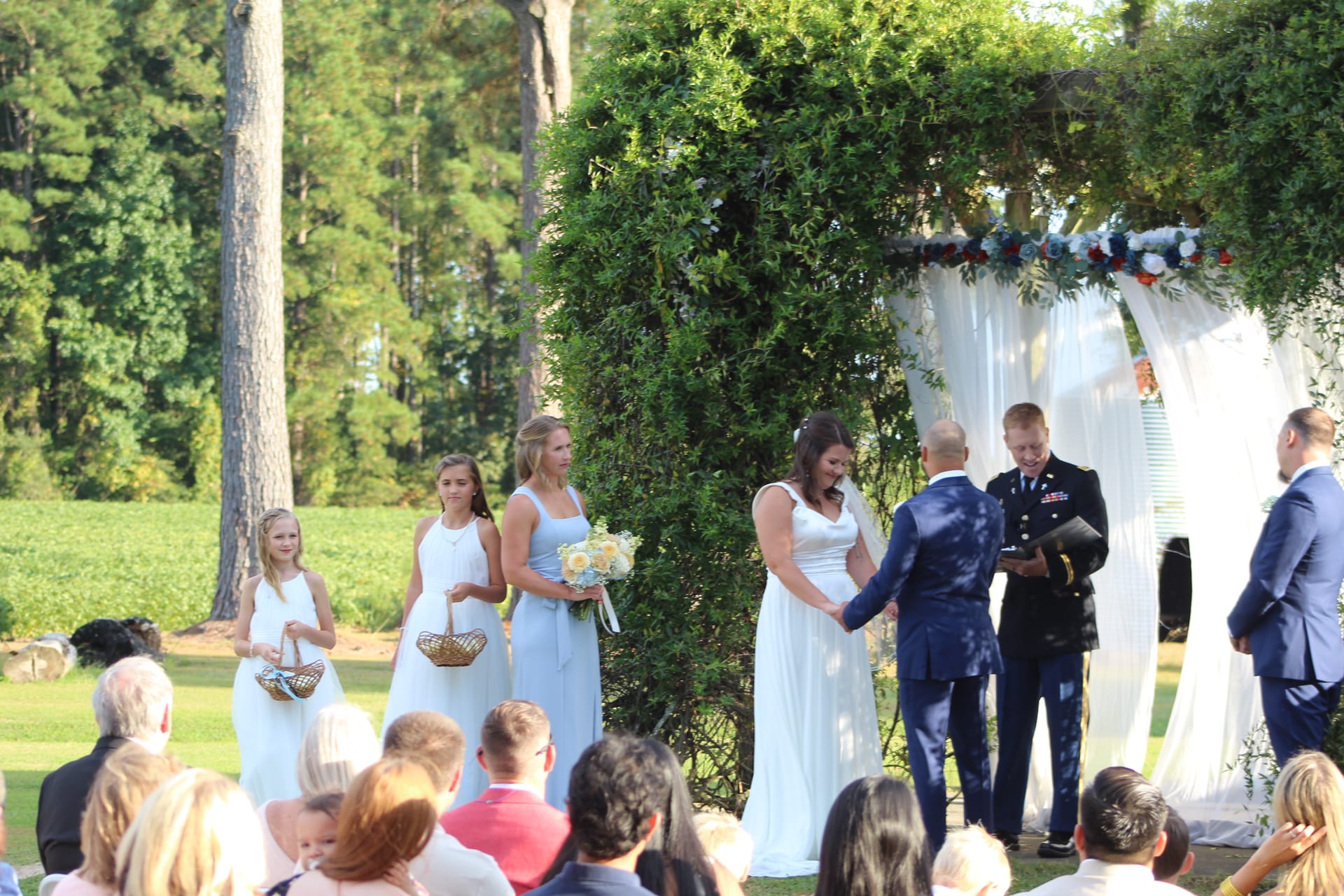Bride and groom exchange vows beneath a lush green floral arch draped in white fabric on the ceremony lawn at Scarborough House, flanked by bridesmaids in light blue and a military officiant.