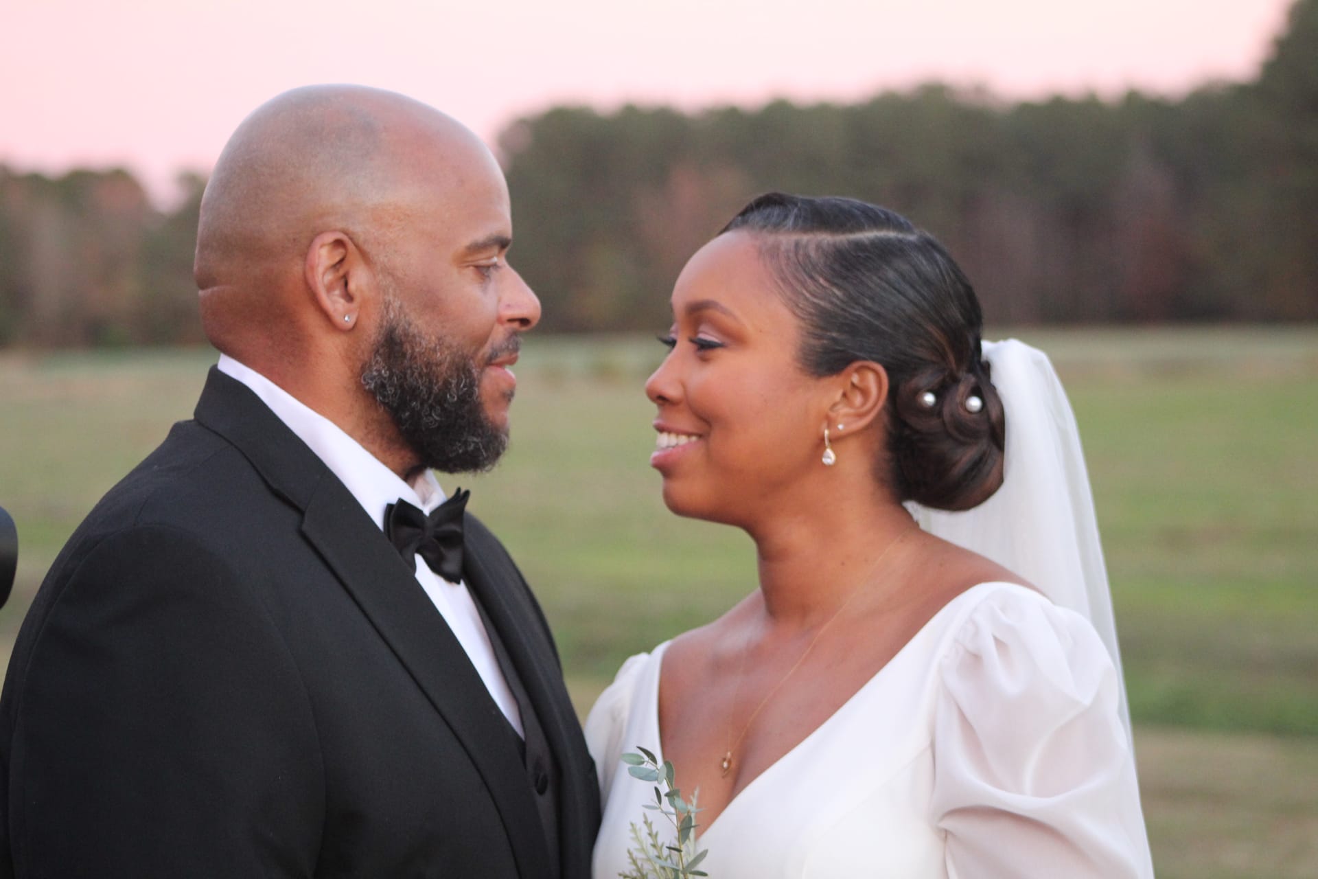 Bride in a white gown and groom in a black tuxedo share an intimate gaze during golden hour against open farmland fields and a soft pink sunset sky at Scarborough House.