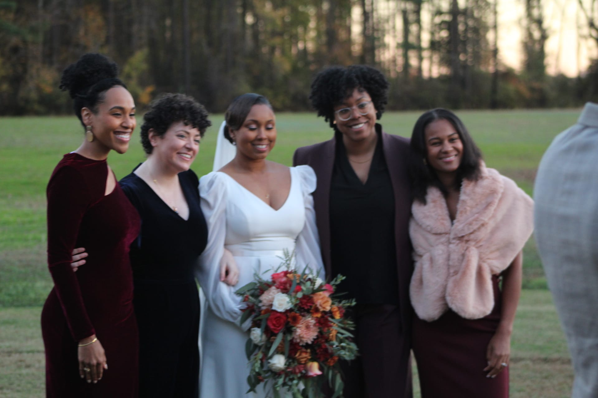A bride in a white gown holding a colorful autumn bouquet poses and laughs with four guests on the ceremony lawn at Scarborough House during golden-hour light with trees in the background.