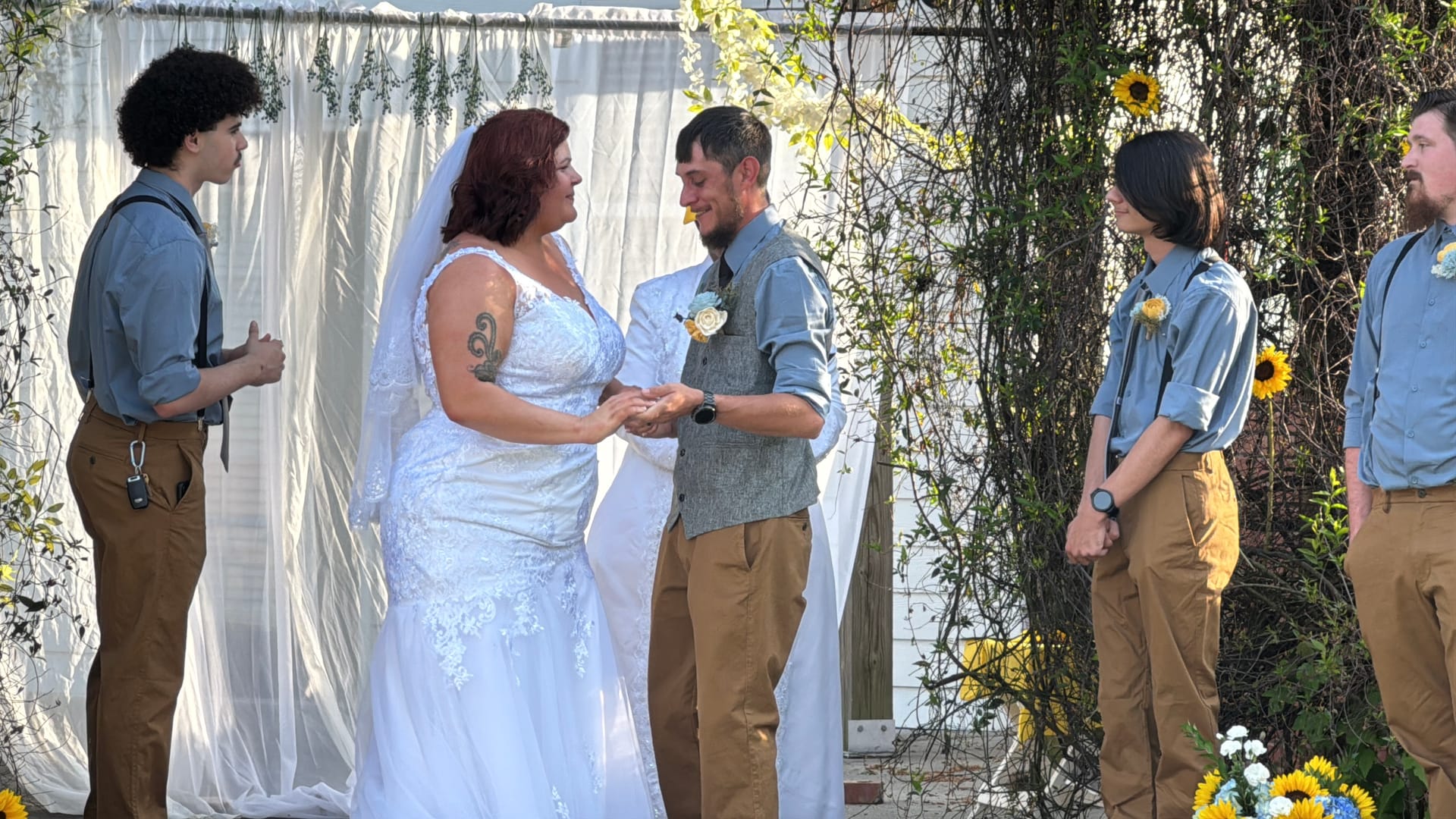 Bride in white lace gown and groom in grey vest exchange rings at an outdoor ceremony framed by draped fabric and sunflowers with groomsmen looking on.
