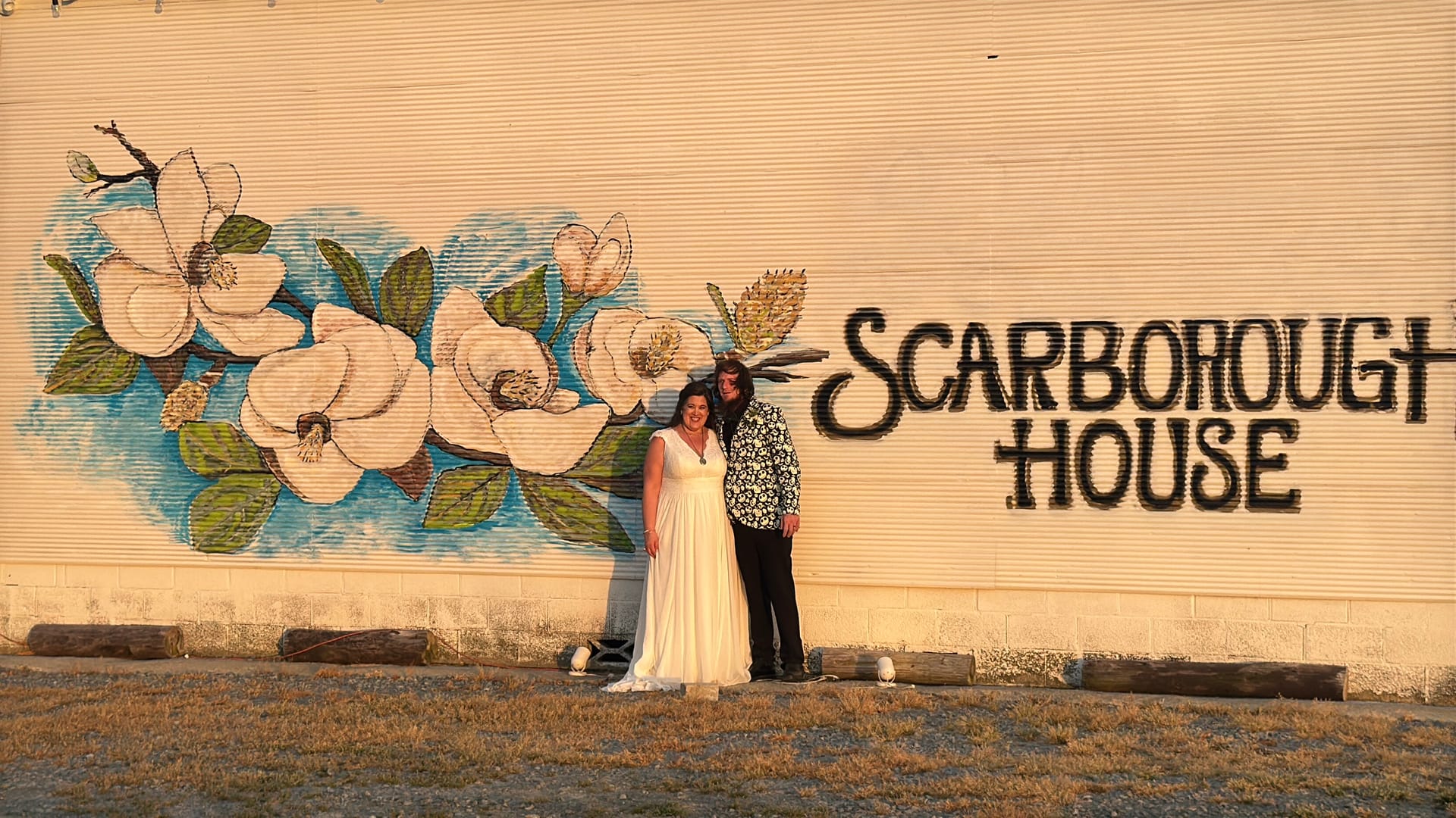 A bride in a white gown and her partner in a floral blazer pose together in front of the Scarborough House magnolia mural during golden hour.