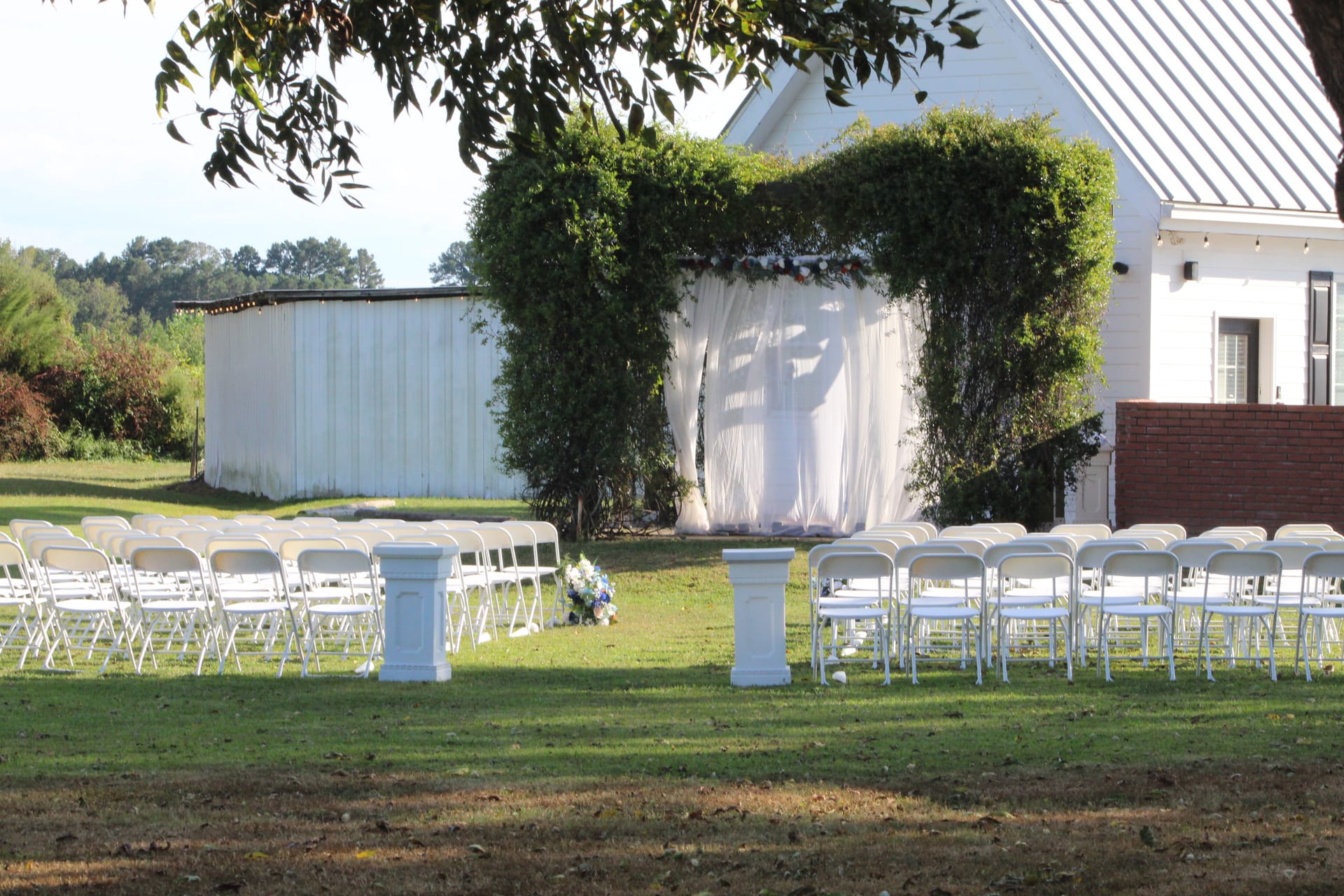 Outdoor wedding ceremony setup at Scarborough House featuring rows of white folding chairs flanking a central aisle leading to a lush ivy-covered arch draped with white fabric and floral accents beside the historic white manor.