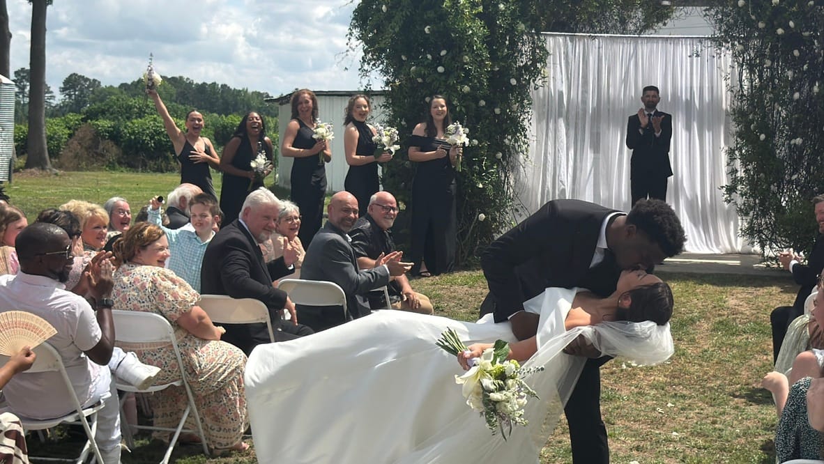 Groom dips and kisses his bride at the end of their outdoor wedding ceremony as bridesmaids and guests cheer and applaud on the sunlit lawn.
