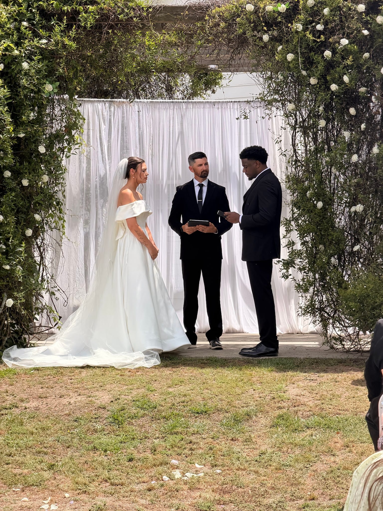 Bride in an off-shoulder ball gown and groom in a black suit exchange vows before a white draped backdrop framed by a lush floral arch on the ceremony lawn at Scarborough House.