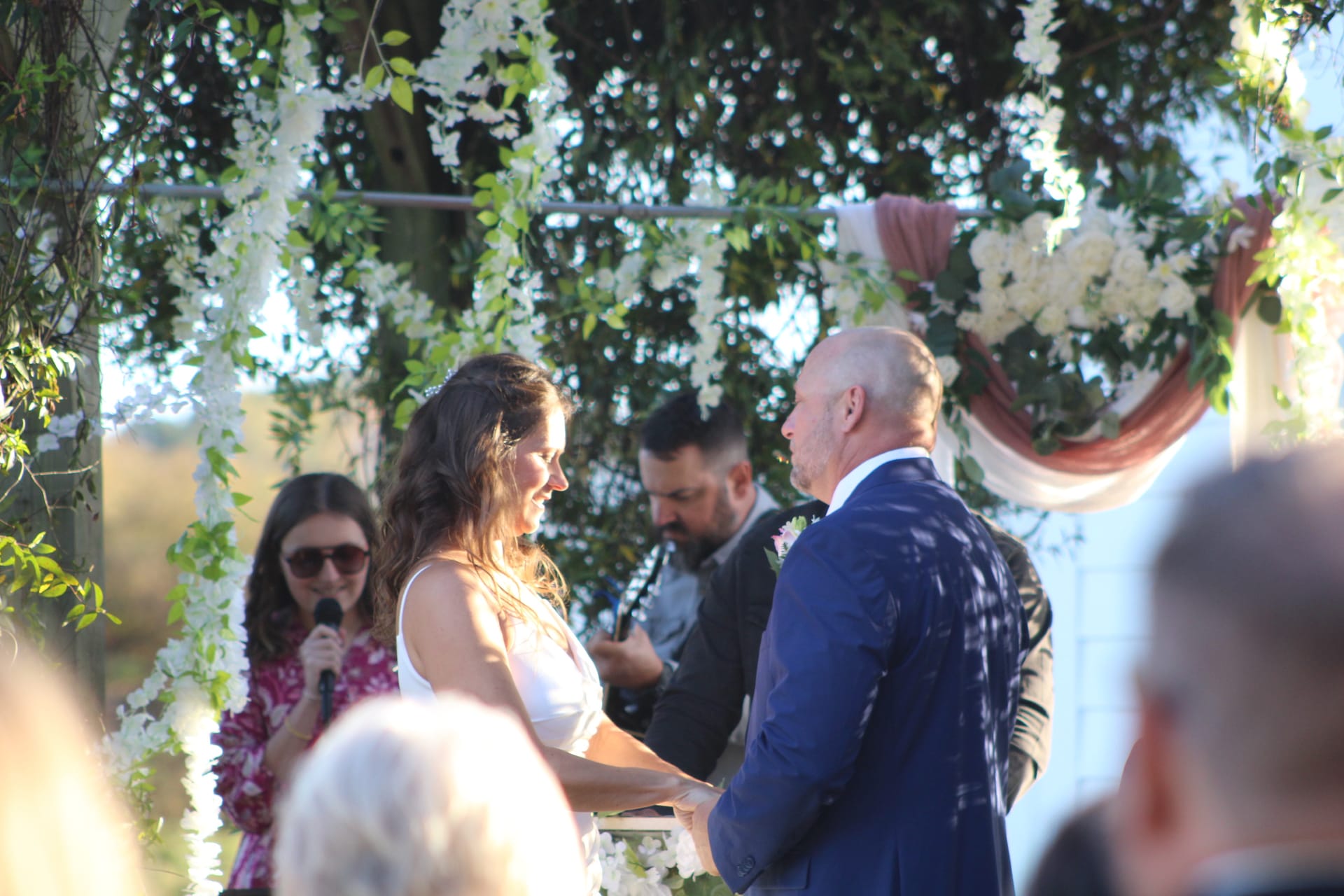 Bride and groom holding hands and smiling at each other during their outdoor wedding ceremony beneath a floral arch adorned with white blooms and greenery at Scarborough House.