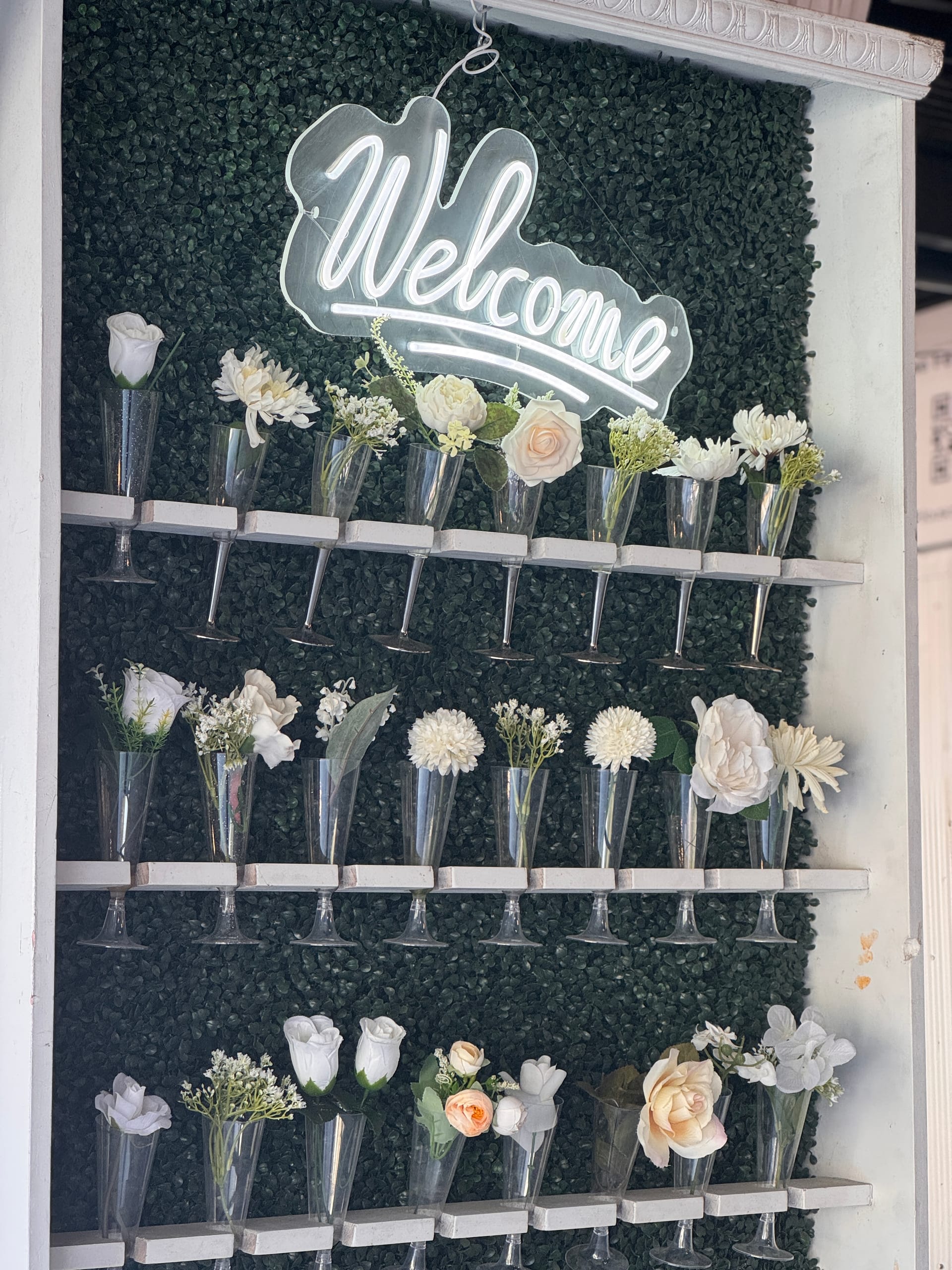 A decorative welcome wall at Scarborough House featuring rows of champagne flute vases filled with white and blush flowers mounted on white shelves against a boxwood greenery backdrop with a glowing neon Welcome sign.