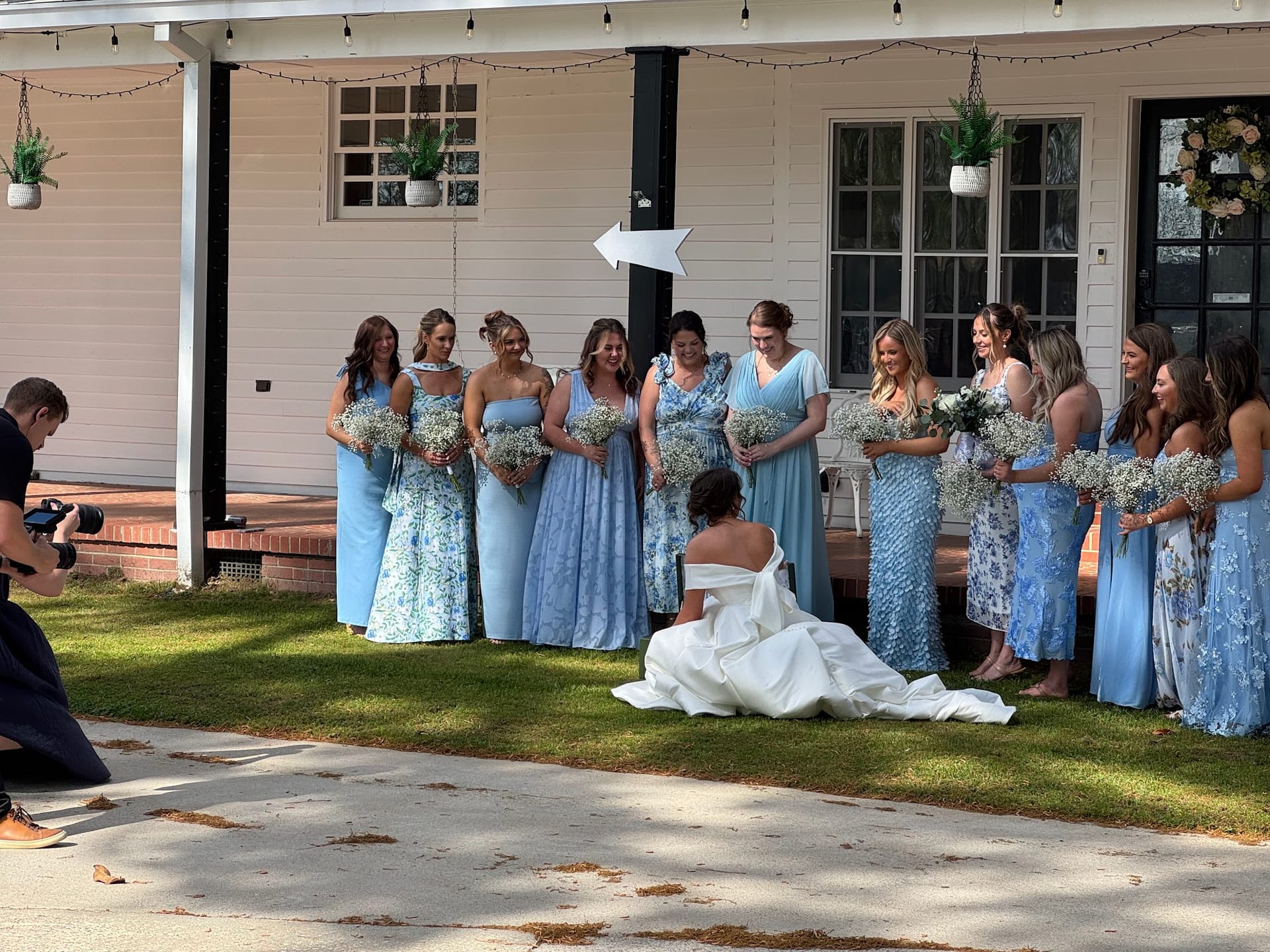 Bridesmaids in shades of blue holding white bouquets smile and laugh as the bride sits on the lawn in her white gown during a wedding party photo session at Scarborough House.