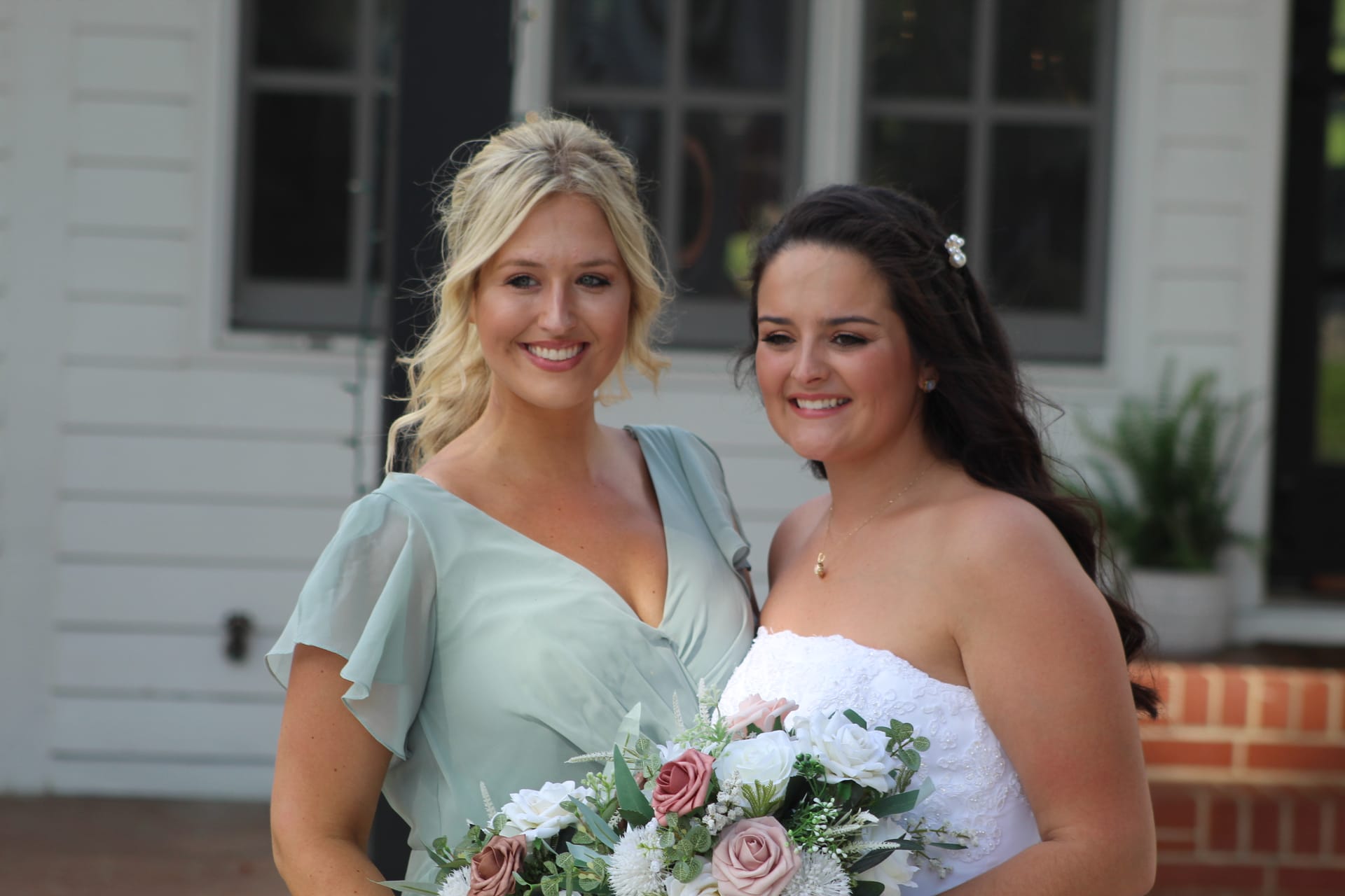 Bride in a strapless white lace gown holding a pink and white bouquet poses with a bridesmaid in a sage green dress in front of the historic 1821 Scarborough House manor.