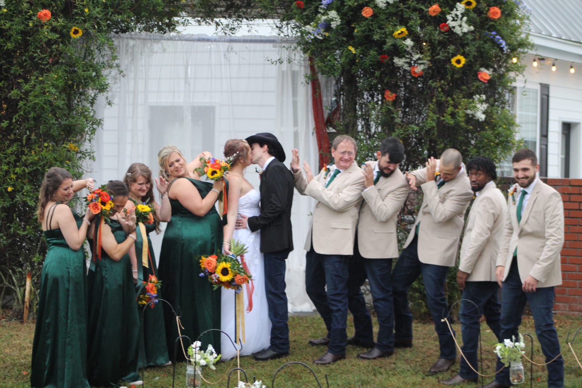 Bride and groom share their first kiss under a lush floral arch on the ceremony lawn at Scarborough House while bridesmaids in emerald gowns and groomsmen in tan blazers cheer around them.