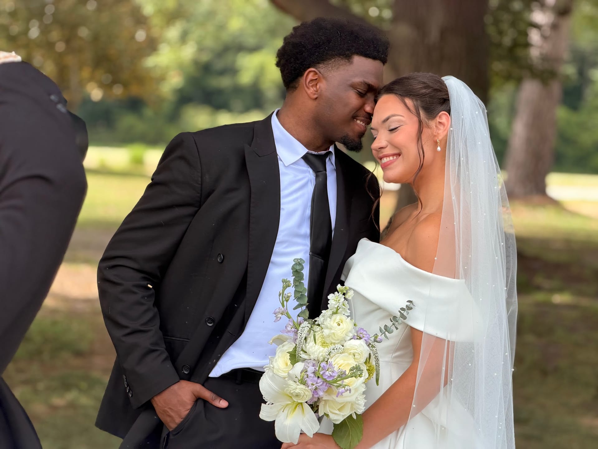 Bride and groom share a tender forehead-touch moment surrounded by oak trees on the estate grounds, the bride holding a bouquet of white lilies and lavender blooms.