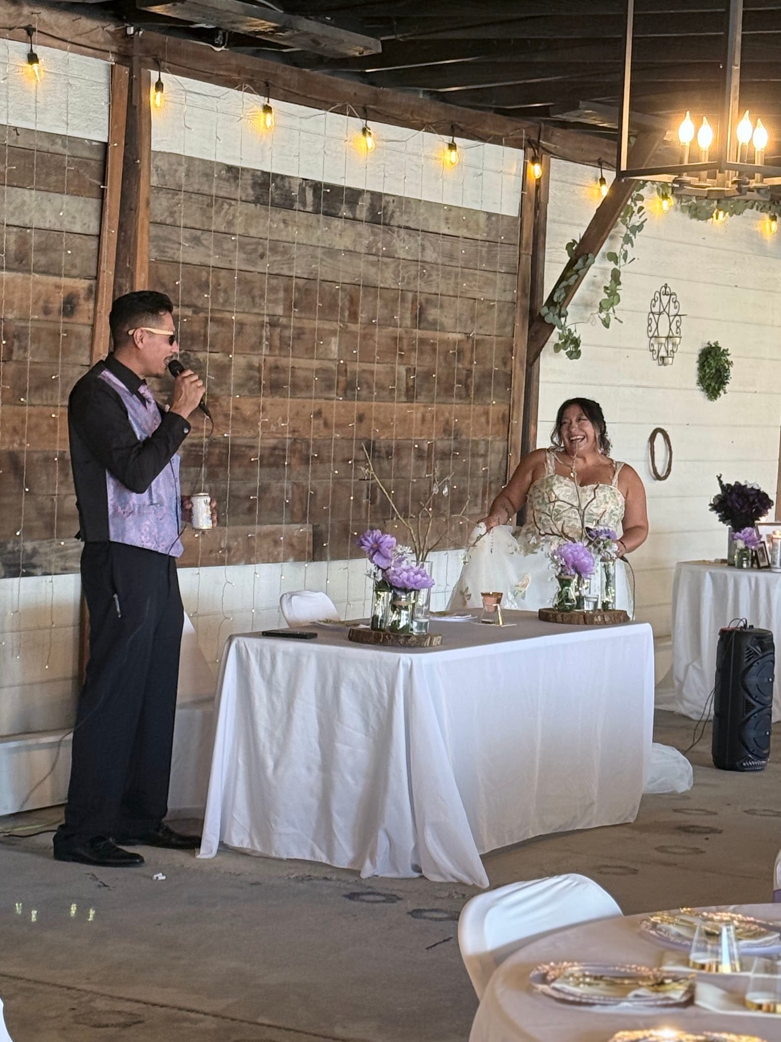 Groom speaks into a microphone while delivering a toast as the laughing bride sits at the sweetheart table adorned with purple floral arrangements inside a rustic barn venue strung with warm Edison lights.