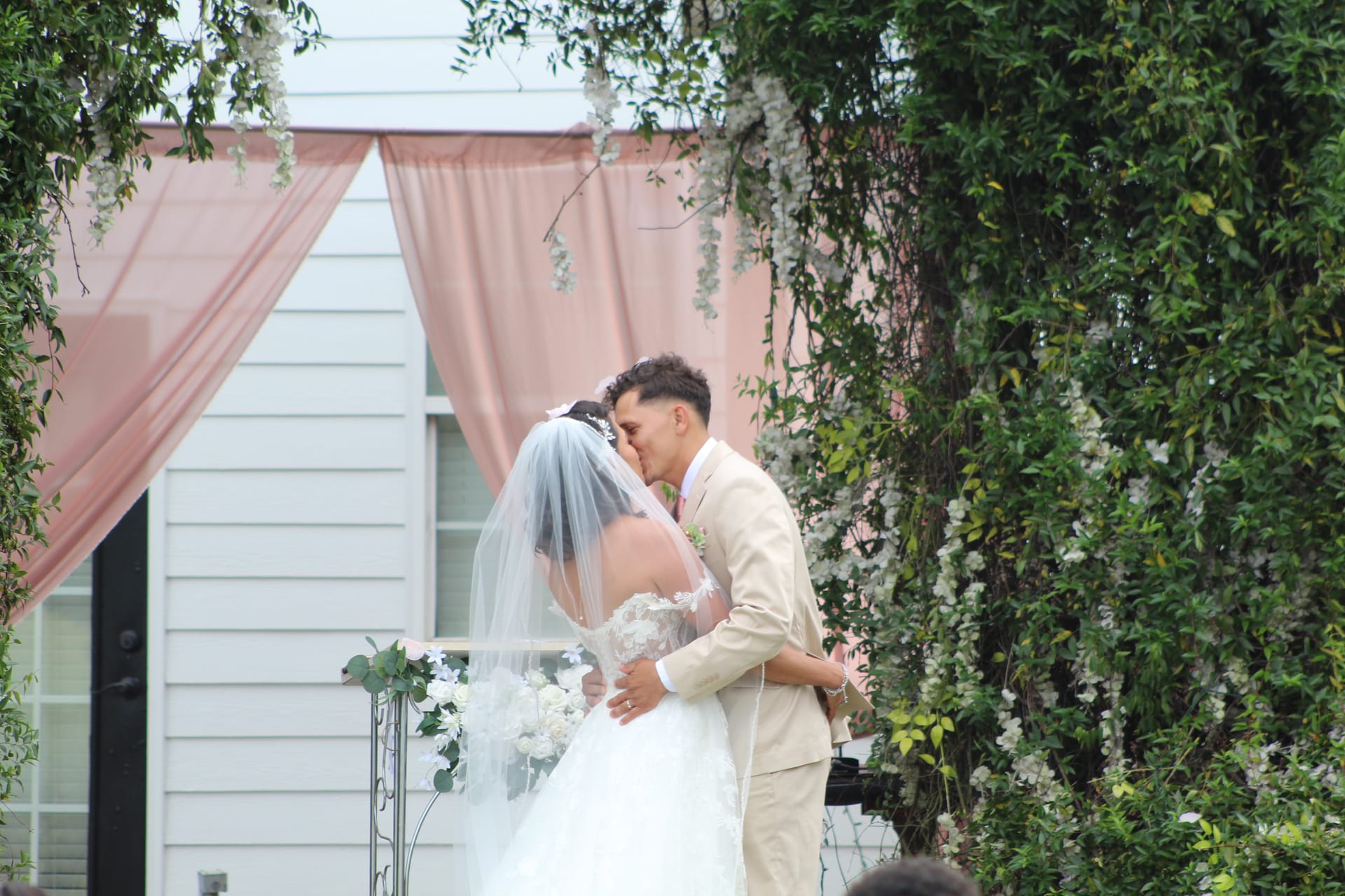 Bride in a lace gown and veil shares her first kiss with the groom in a tan suit beneath a blush-draped arch surrounded by cascading white florals and lush greenery at Scarborough House.