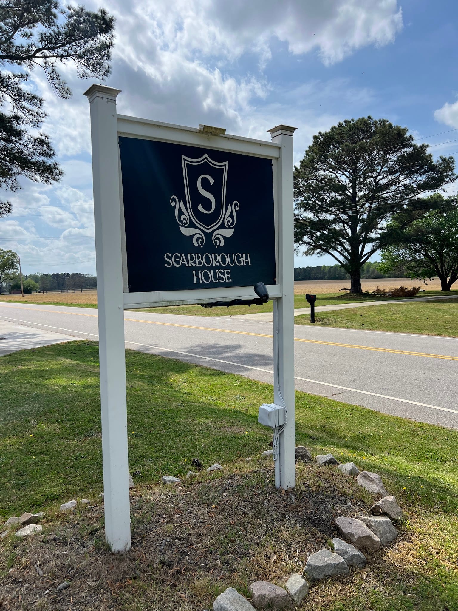 Scarborough House estate wedding venue sign with ornate crest logo mounted on white posts along a rural road flanked by open fields and mature trees.