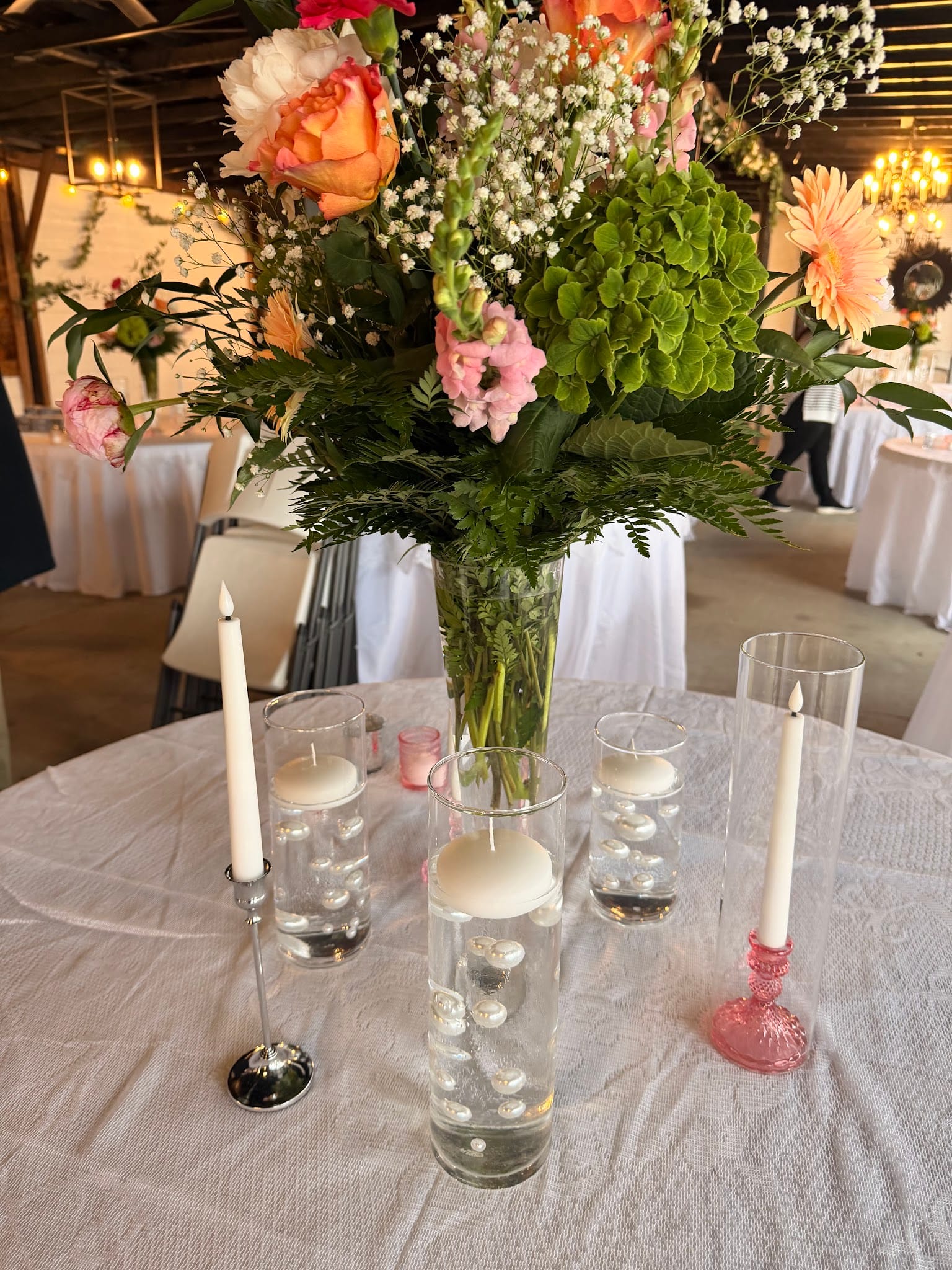 Elegant wedding reception table centerpiece featuring a tall glass vase with orange roses, pink snapdragons, green hydrangeas and baby's breath, surrounded by floating candle cylinders with decorative stones and pink taper candles in a warmly lit barn venue.