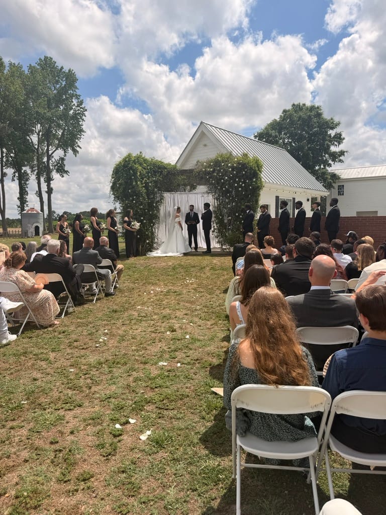 Outdoor wedding ceremony on the lawn at Scarborough House with guests seated in white chairs facing the couple at a floral archway in front of a white historic chapel.