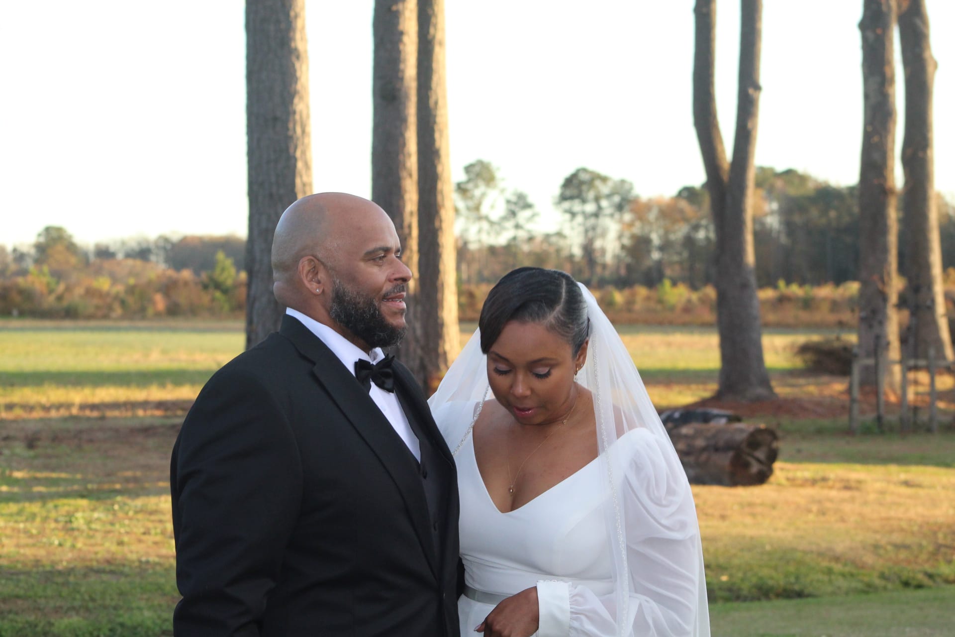 Bride in a white long-sleeve gown and veil stands beside her tuxedo-clad groom as he gazes at her lovingly against the golden-hour farmland fields at Scarborough House.